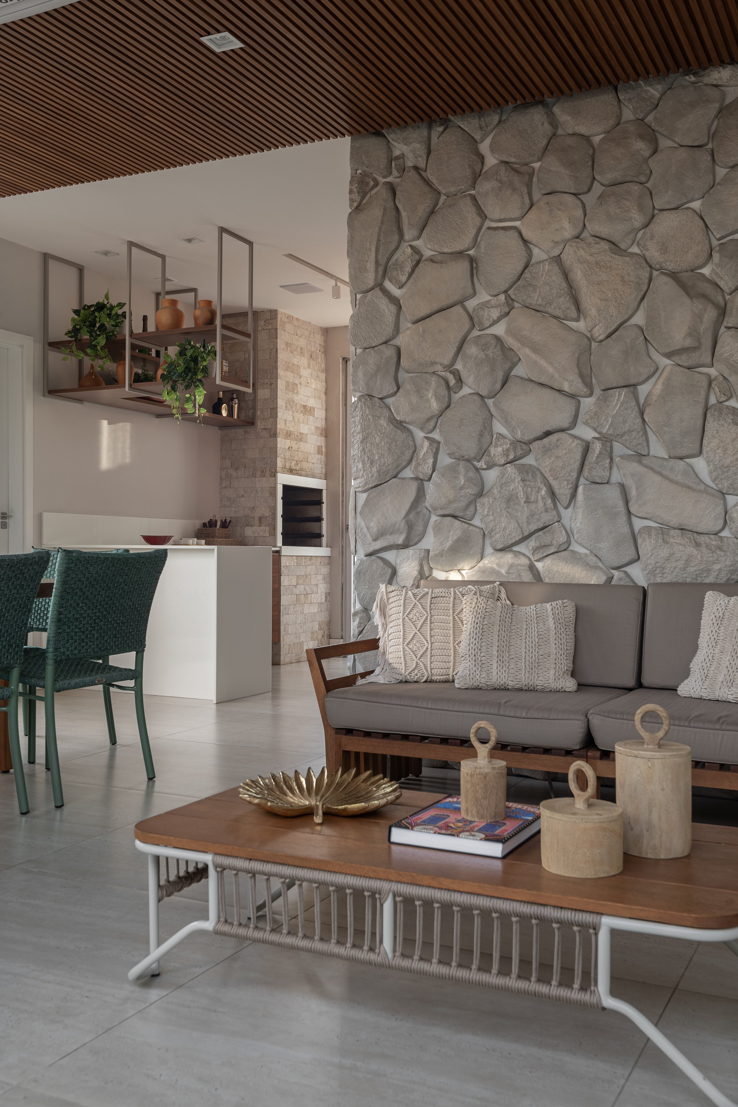 Modern living room interior with a stone accent wall, gray sofa with decorative pillows, wooden coffee table with gold decor, and open kitchen with brick wall and wooden shelving