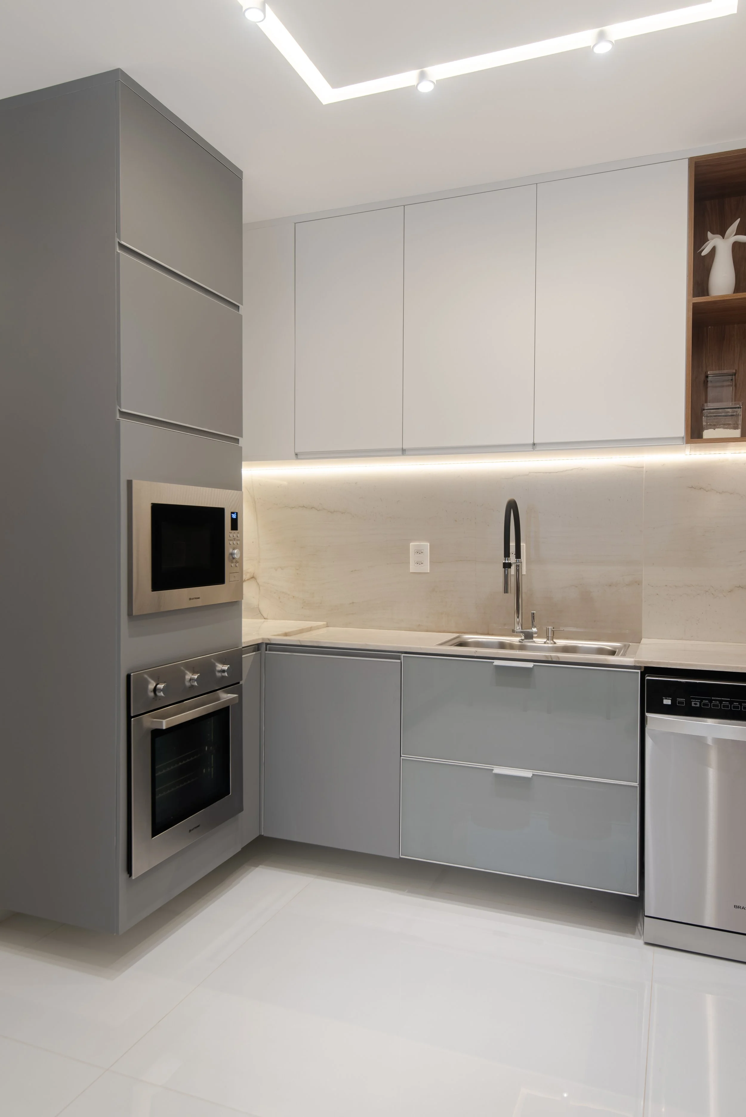 Modern kitchen with gray cabinets, built-in microwave and oven, sink with black faucet, and white backsplash with under-cabinet lighting.