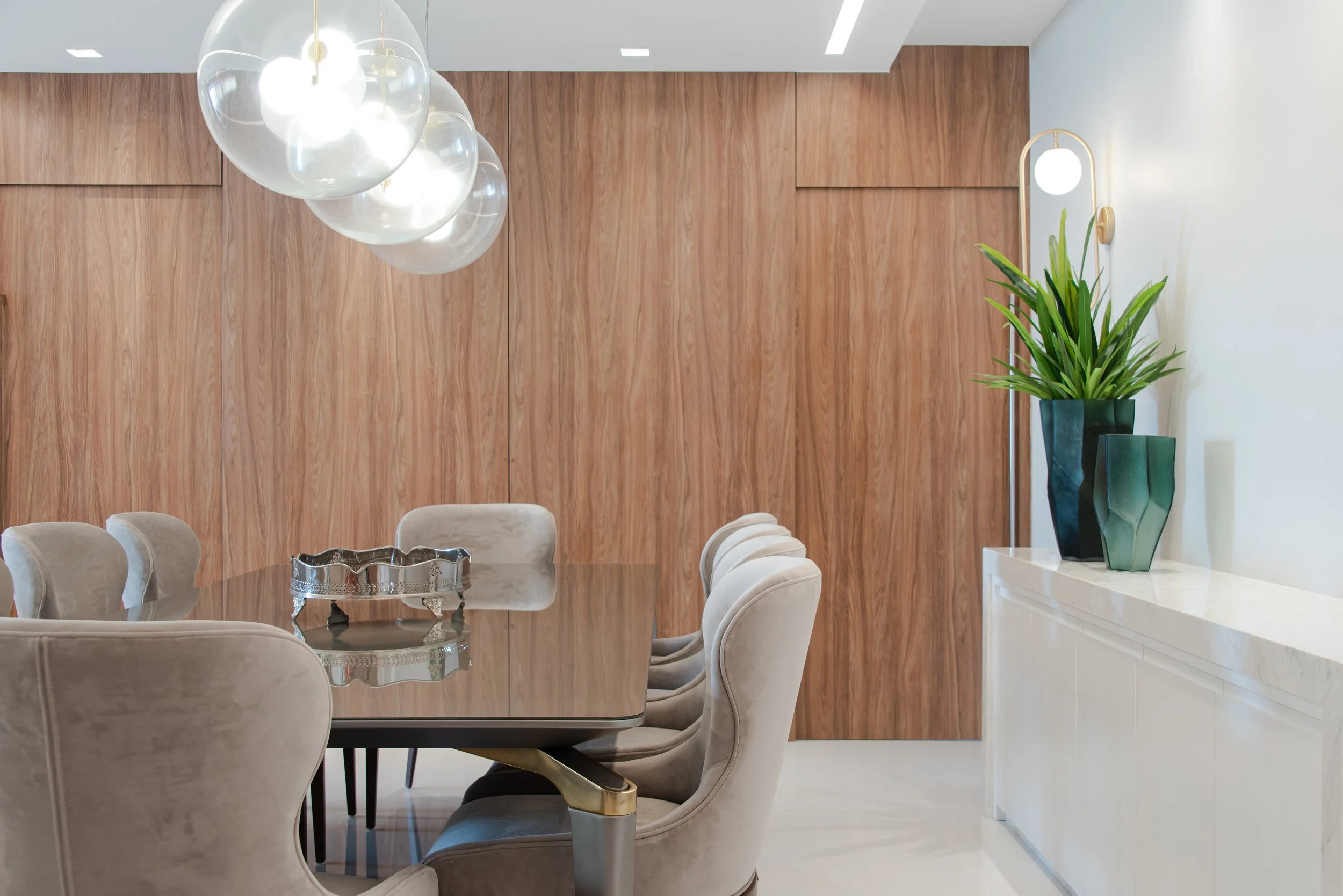 Modern dining room with a wooden wall, a large wooden dining table with beige cushioned chairs, a chandelier with multiple glass globes, and green plants in ceramic vases on a white sideboard.