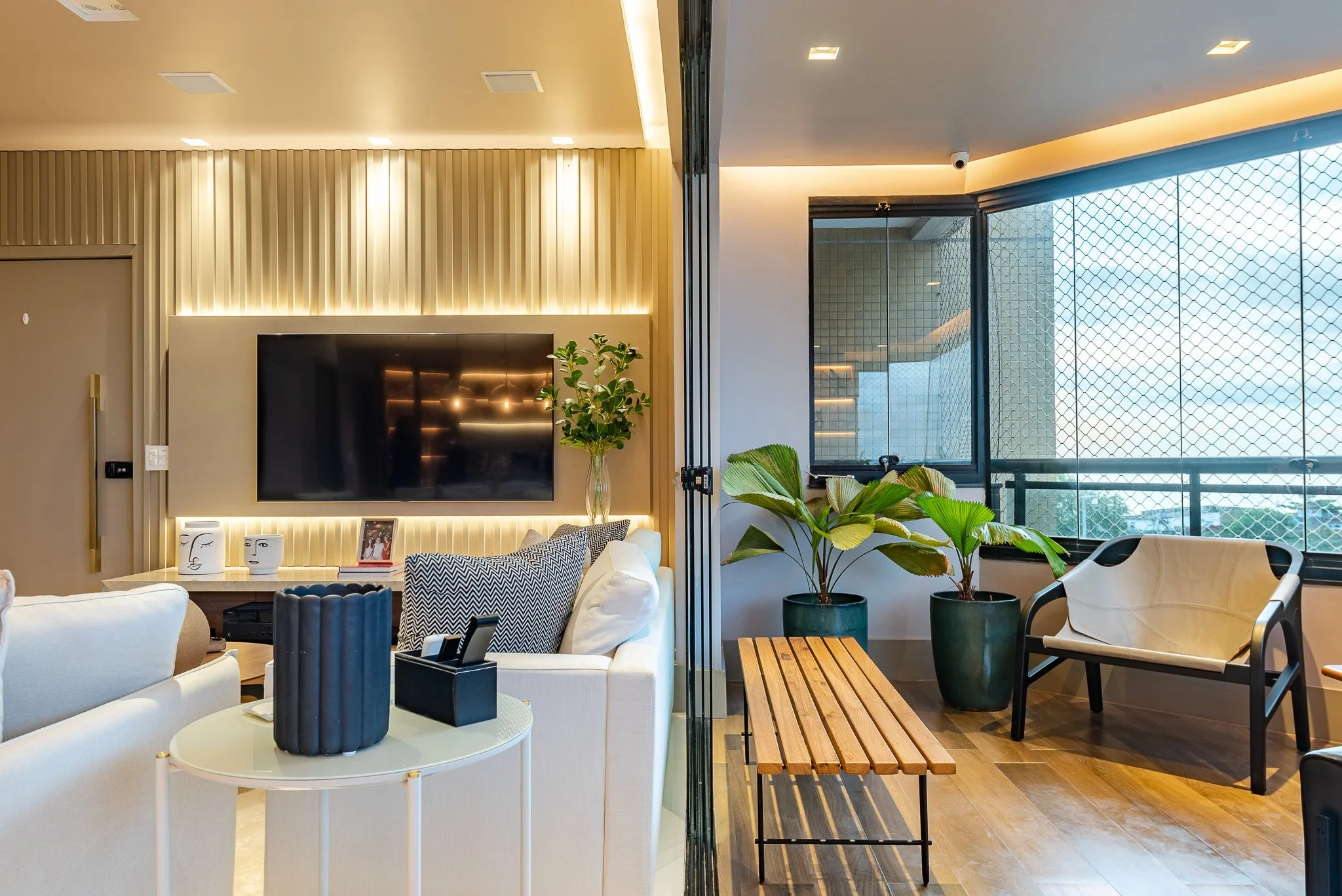 Split view of modern living room with beige and white furniture, large flat-screen TV, decorative vases, framed photo, plants, and a balcony with glass and metal mesh railing
