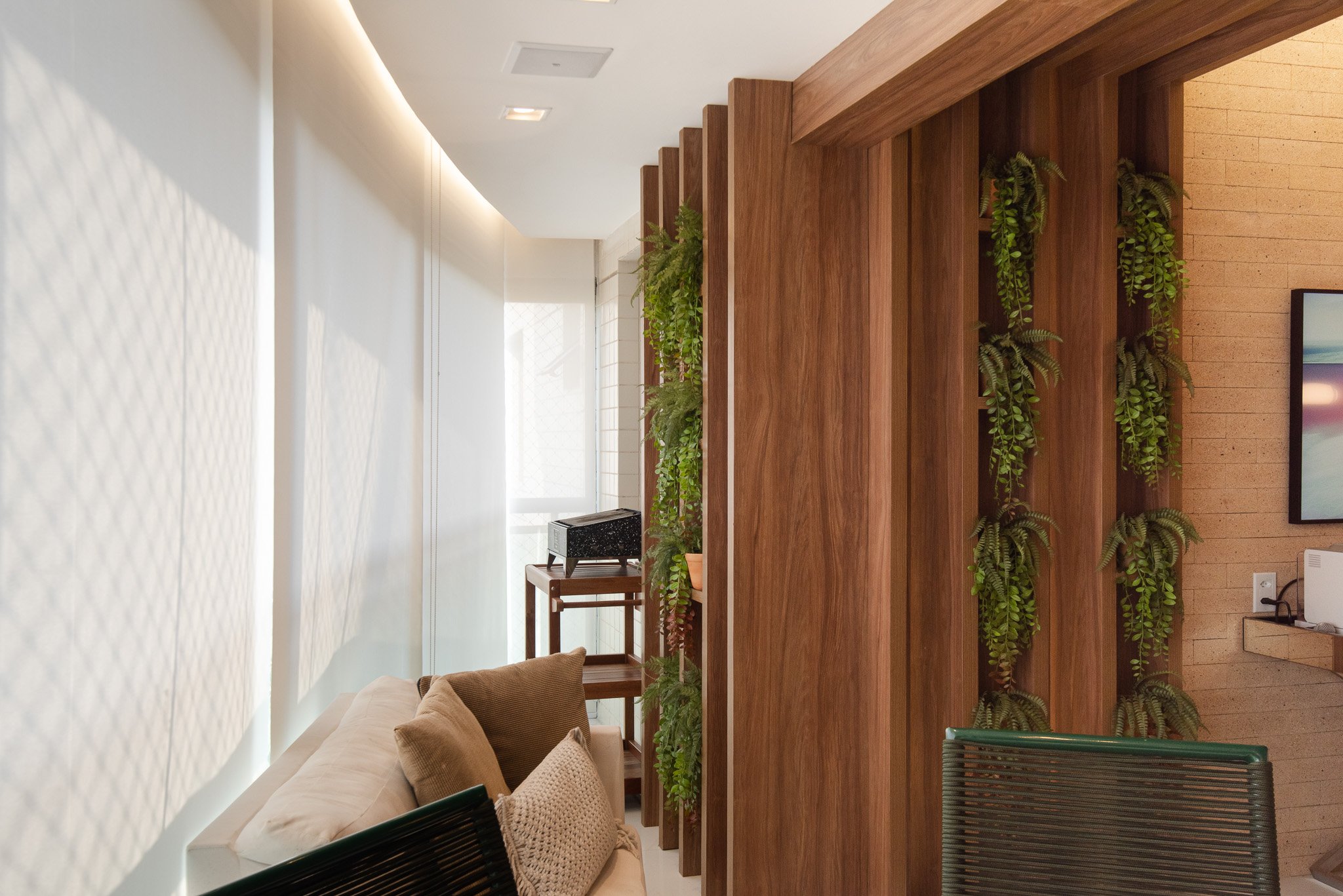 Interior of a room with a beige couch, a wooden partition with green hanging plants, a TV, and a window letting in natural light.