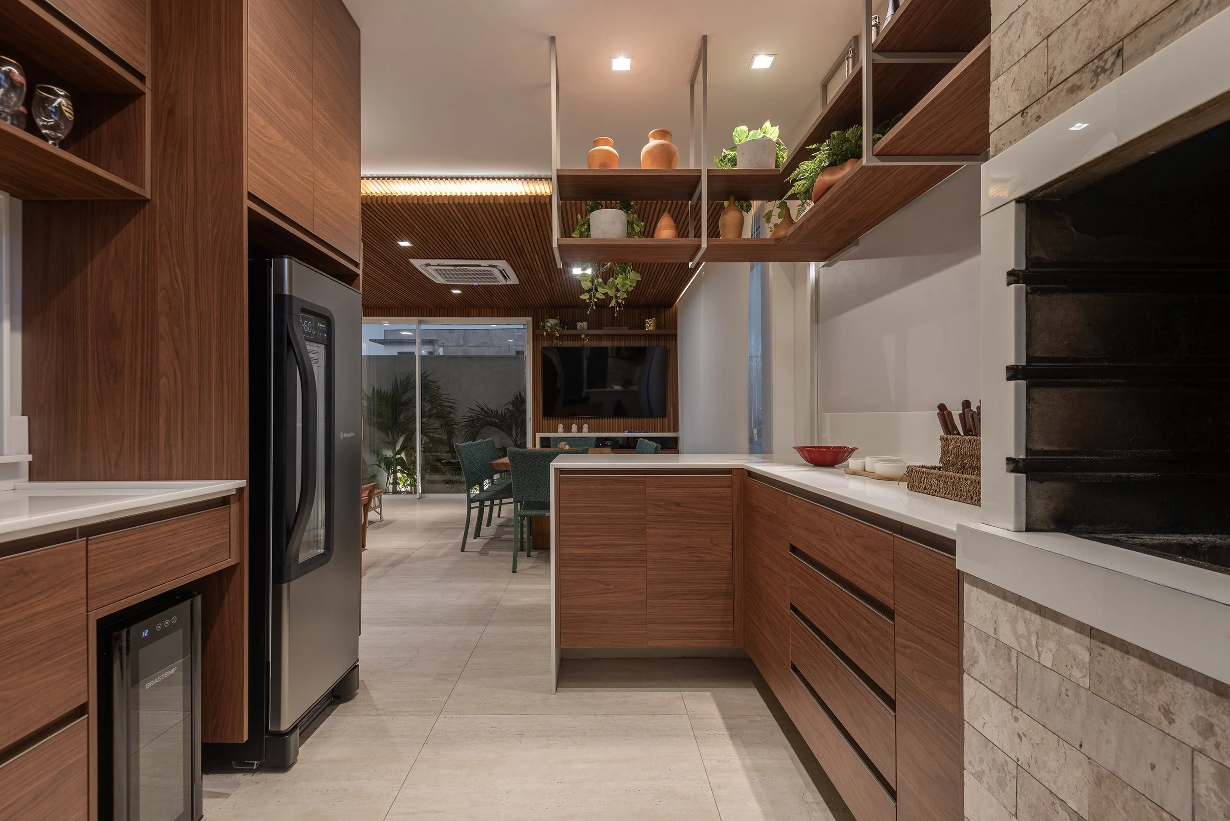Modern kitchen with wooden cabinets, white countertops, a built-in wine fridge, open shelves with plants and pottery, and a view into the dining area with chairs and a wall-mounted TV.