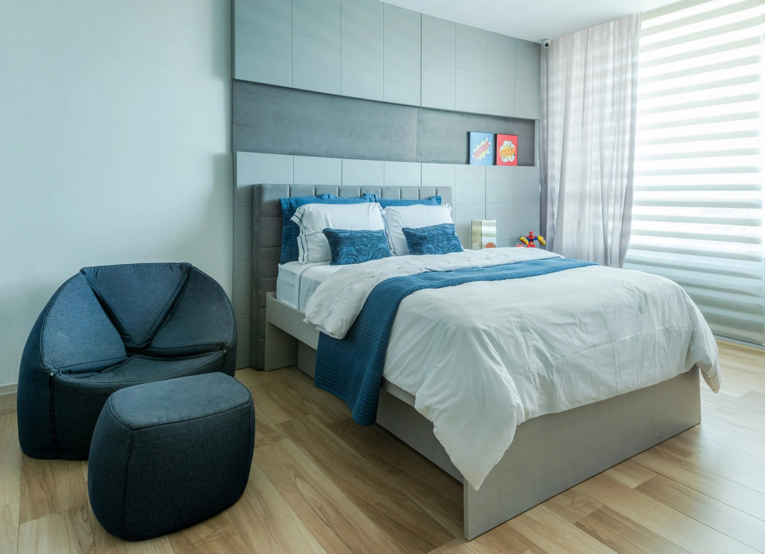 Modern bedroom with a large bed decorated with white and blue bedding, two blue pillows, and a gray headboard. Two small colorful pictures are on the wall above the bed. A black rounded chair with matching ottoman and a window with integral horizonta