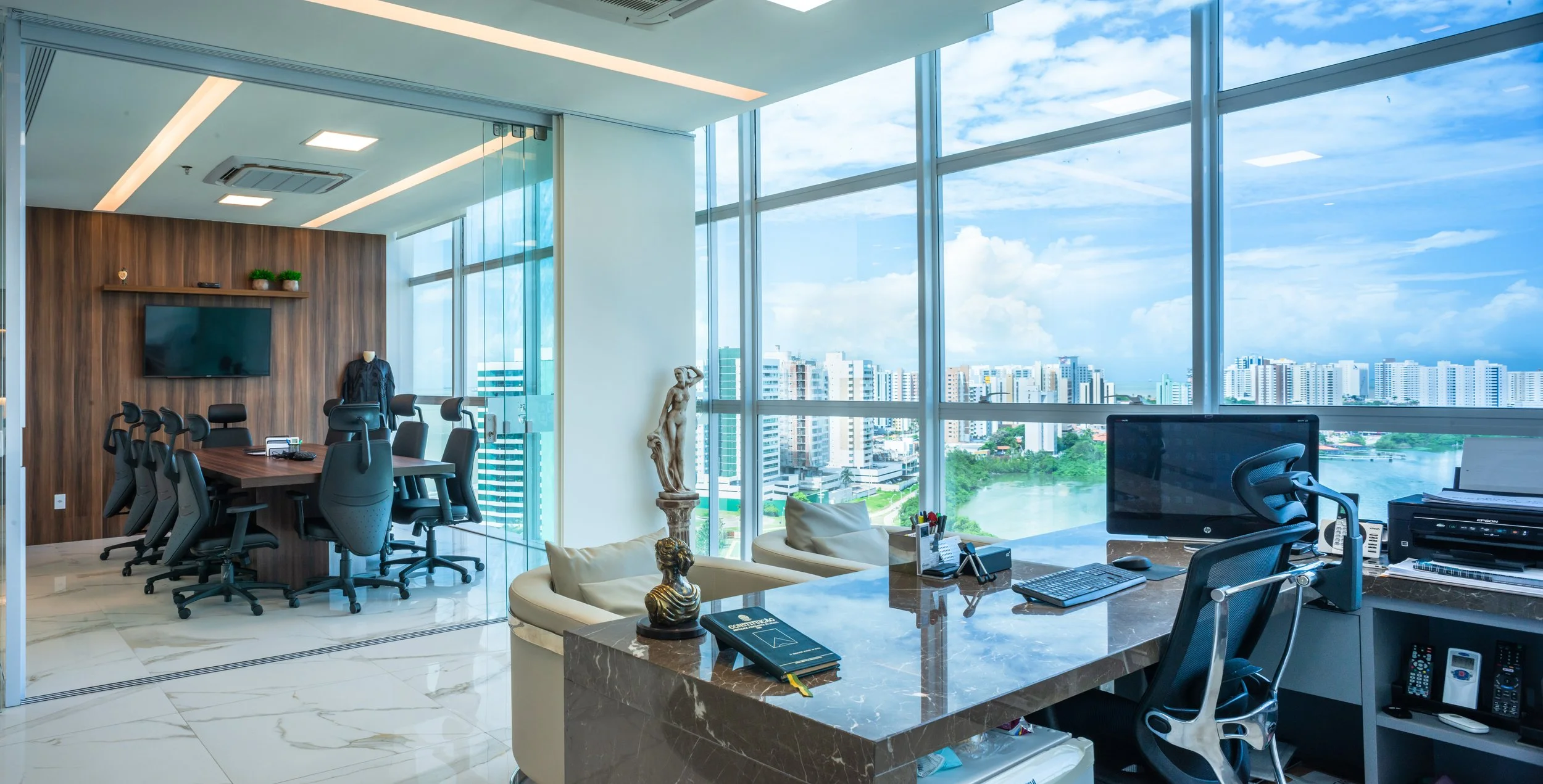 Modern high-rise office with large windows, city skyline view, wooden conference table with multiple chairs, computer desk with monitors and office supplies, decorative statues, and a glass-walled meeting room.