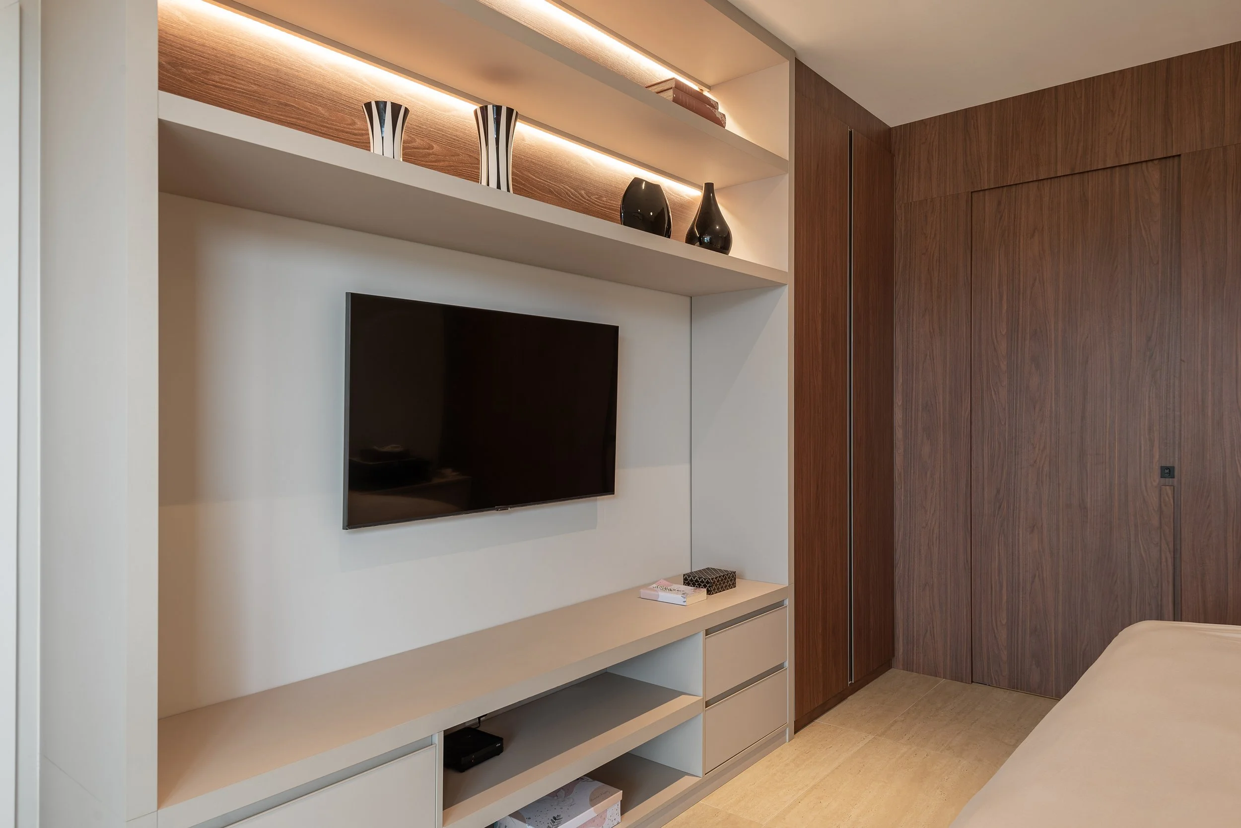 Modern bedroom with a mounted flat-screen TV, white and wood shelving, decorative vases, and a wooden closet door.
