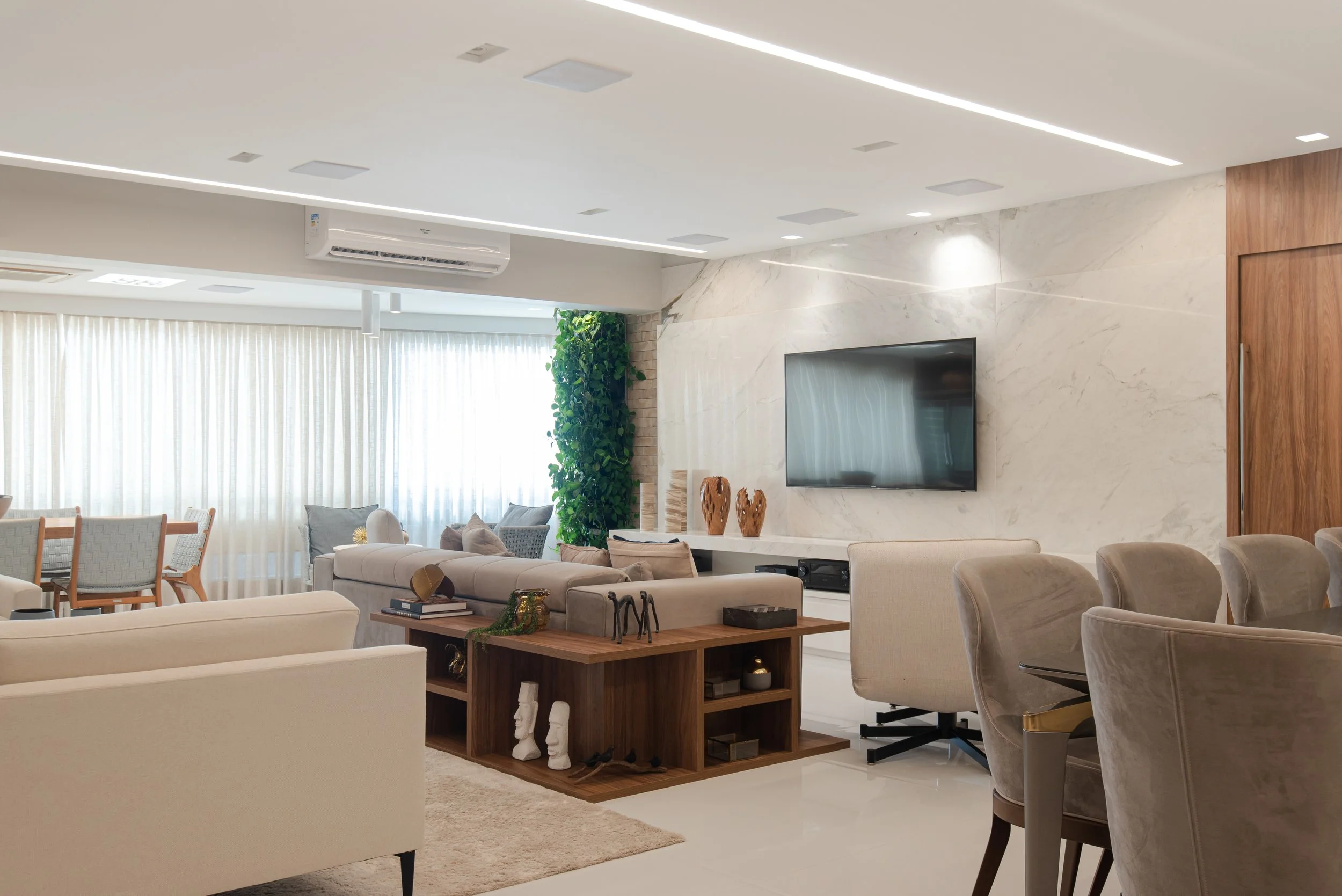 Modern living room with beige sofas, a wall-mounted TV, a wooden partition, and a dining area with chairs near large windows with sheer curtains. Decor includes green wall plants and decorative vases.