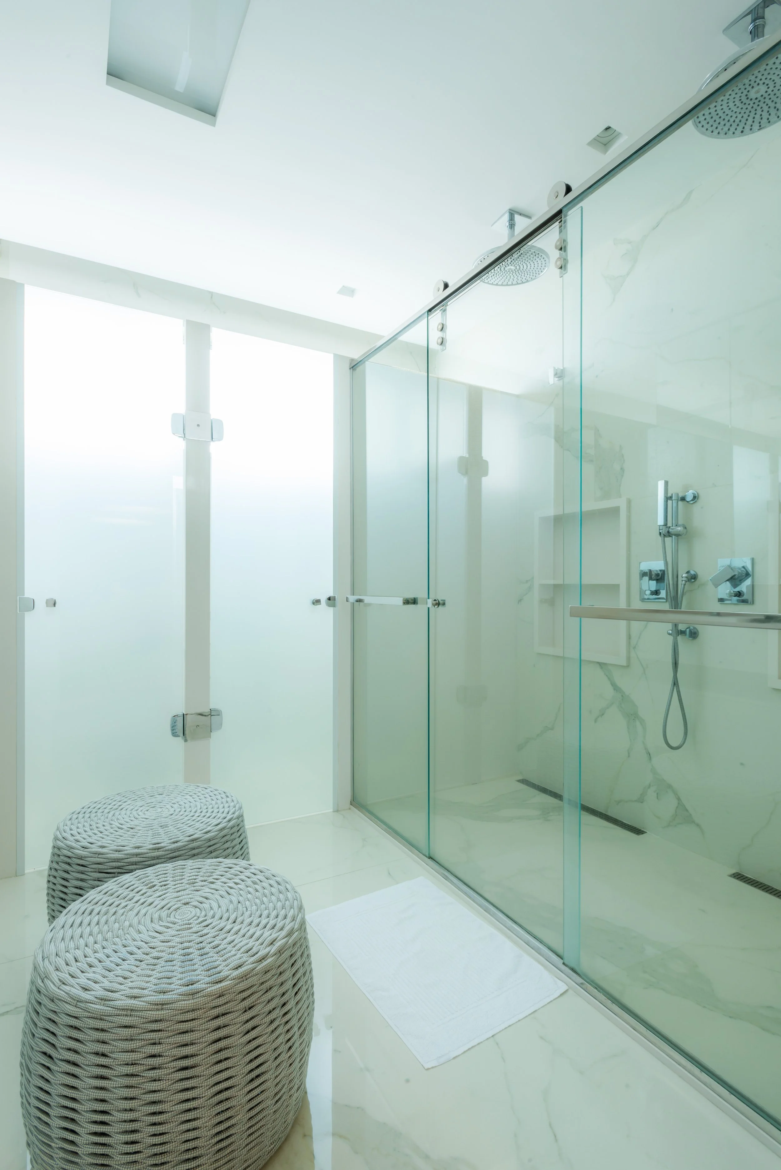 A modern, minimalist bathroom with a glass-enclosed shower, white marble walls, a white bath mat, and two woven round stools.
