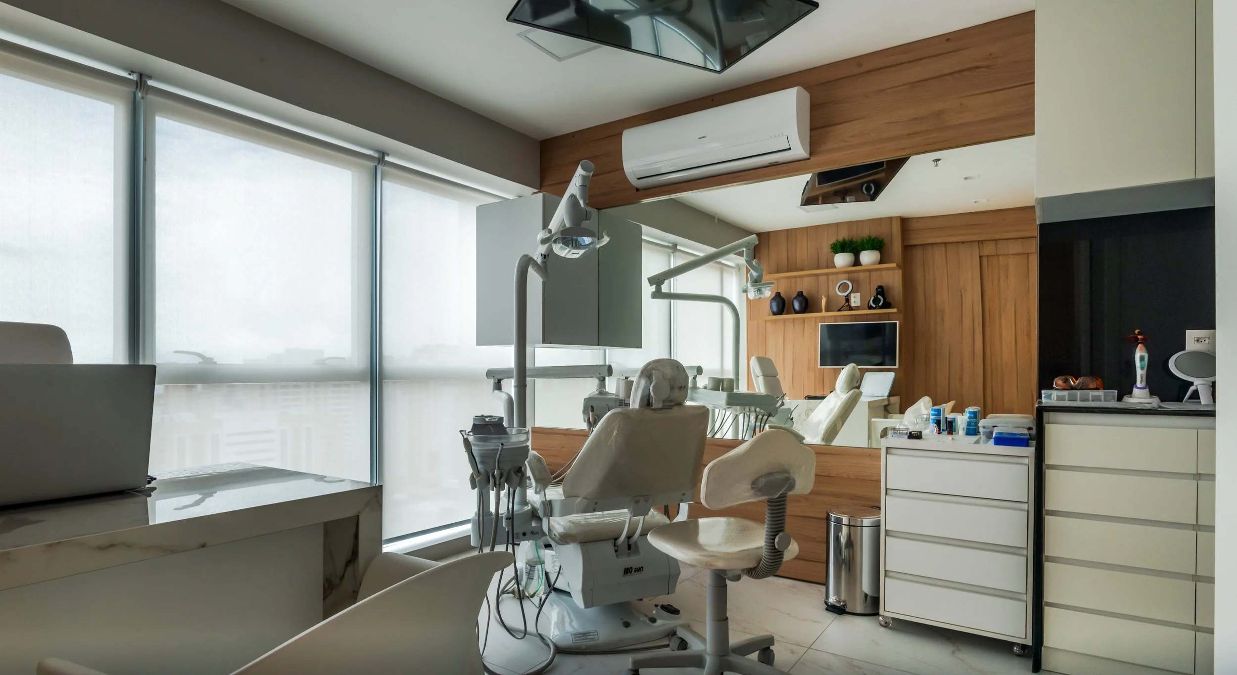 Dental clinic examination room with dental chair, dental tools, and equipment, large windows with blinds, and wooden wall decor.