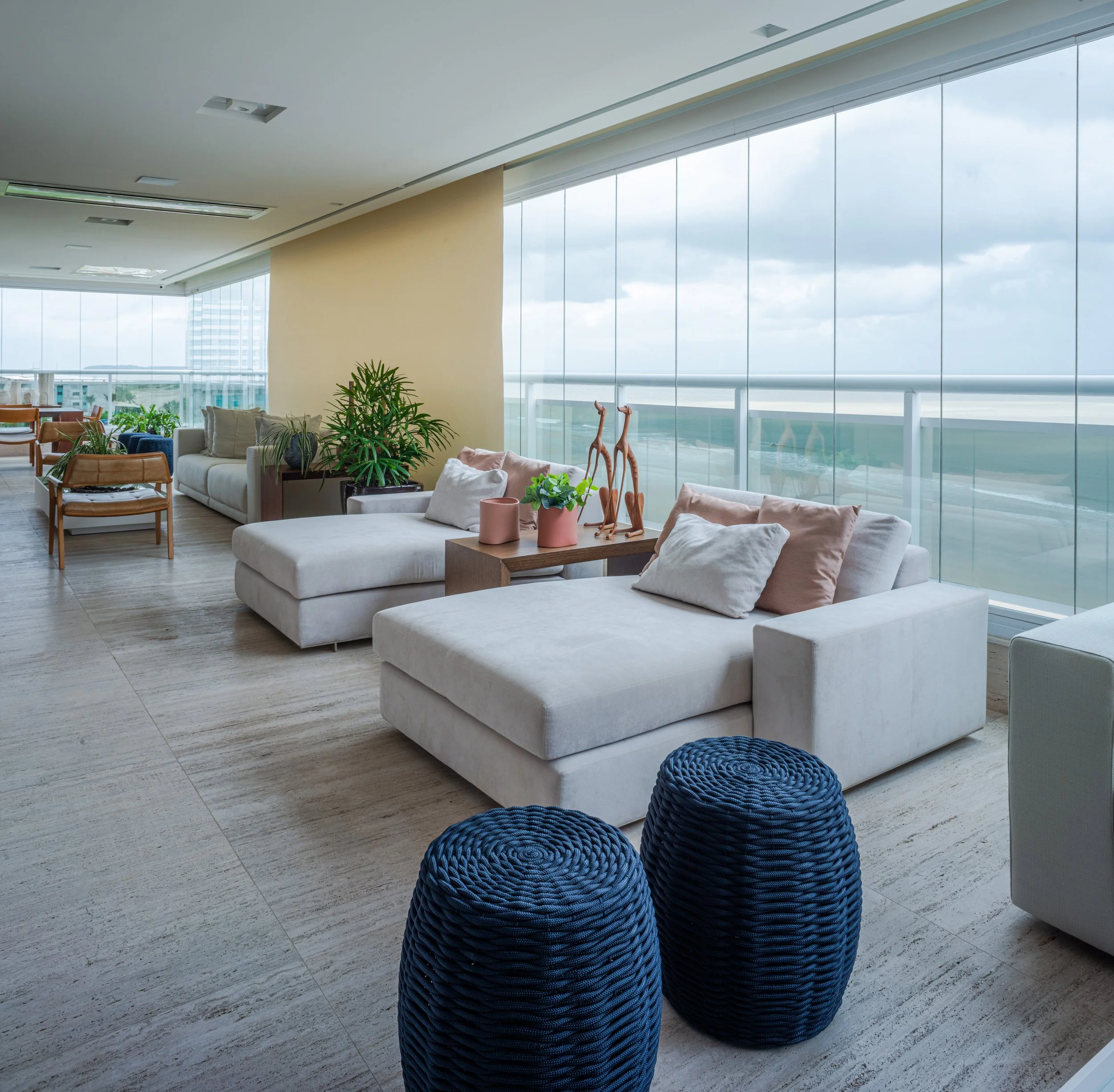 Modern hotel lounge with white sofas, decorative pillows, potted plants, and blue woven ottomans, large windows with a city view, and contemporary decor.