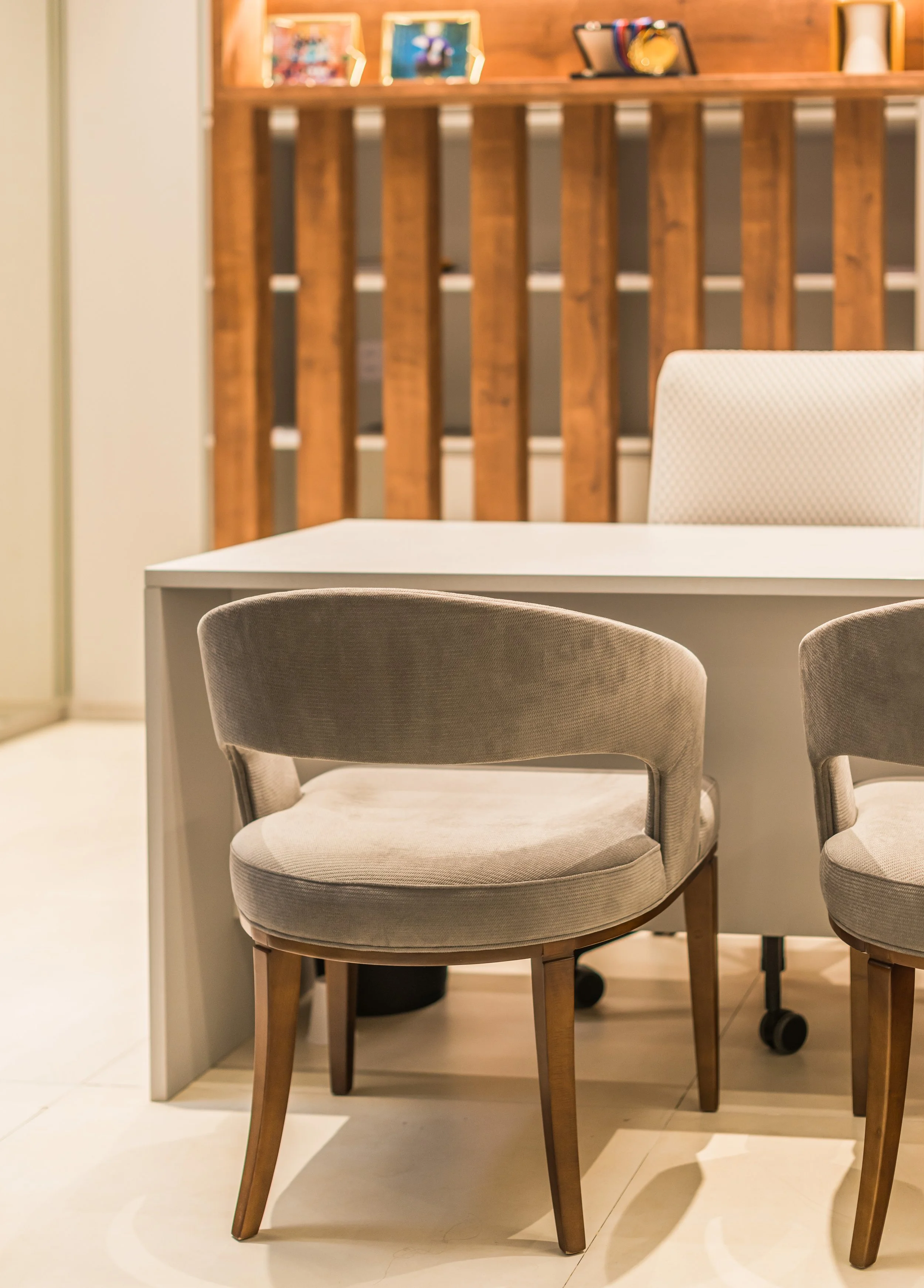 A beige upholstered chair with wooden legs next to a white table in a modern interior. In the background, there is a wooden slat wall with framed photos and decorations on a shelf.