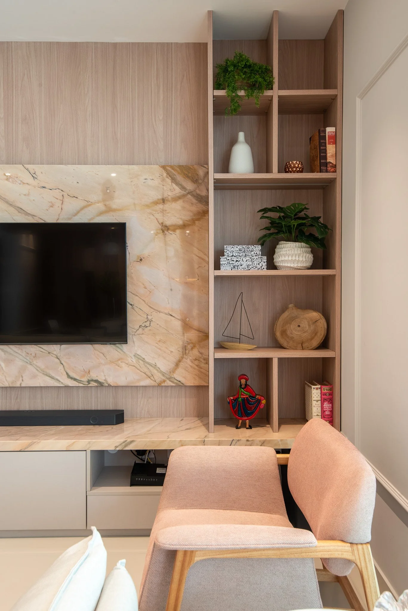 Living room with wooden shelving, marble panel behind a mounted flat-screen TV, decorative items including plants, books, and a small sailboat, beige armchair, and a beige plush chair.