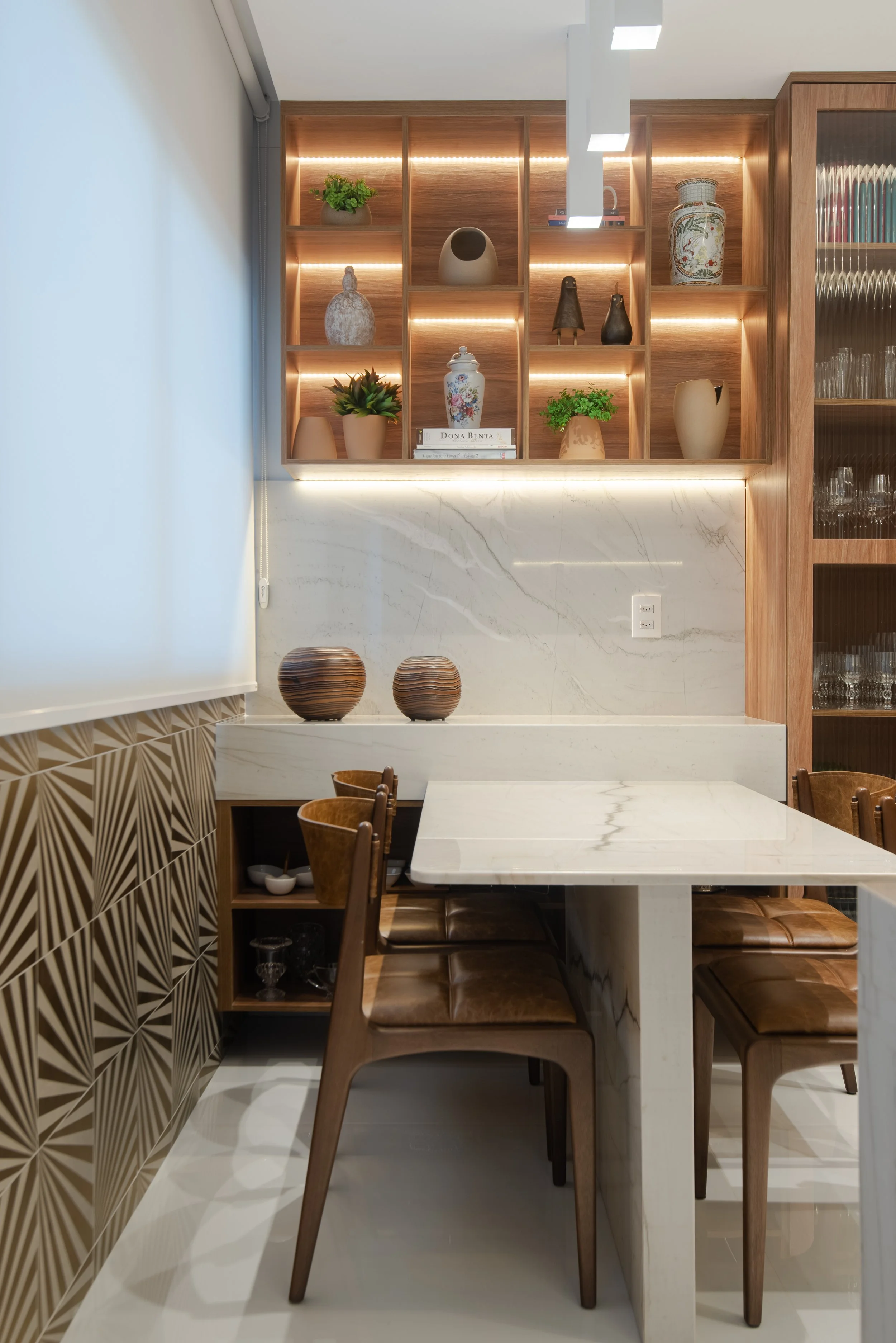 A modern dining area with a wooden and marble design, featuring a built-in open shelving unit with decorative vases, plants, and books, above a marble table and brown leather chairs.