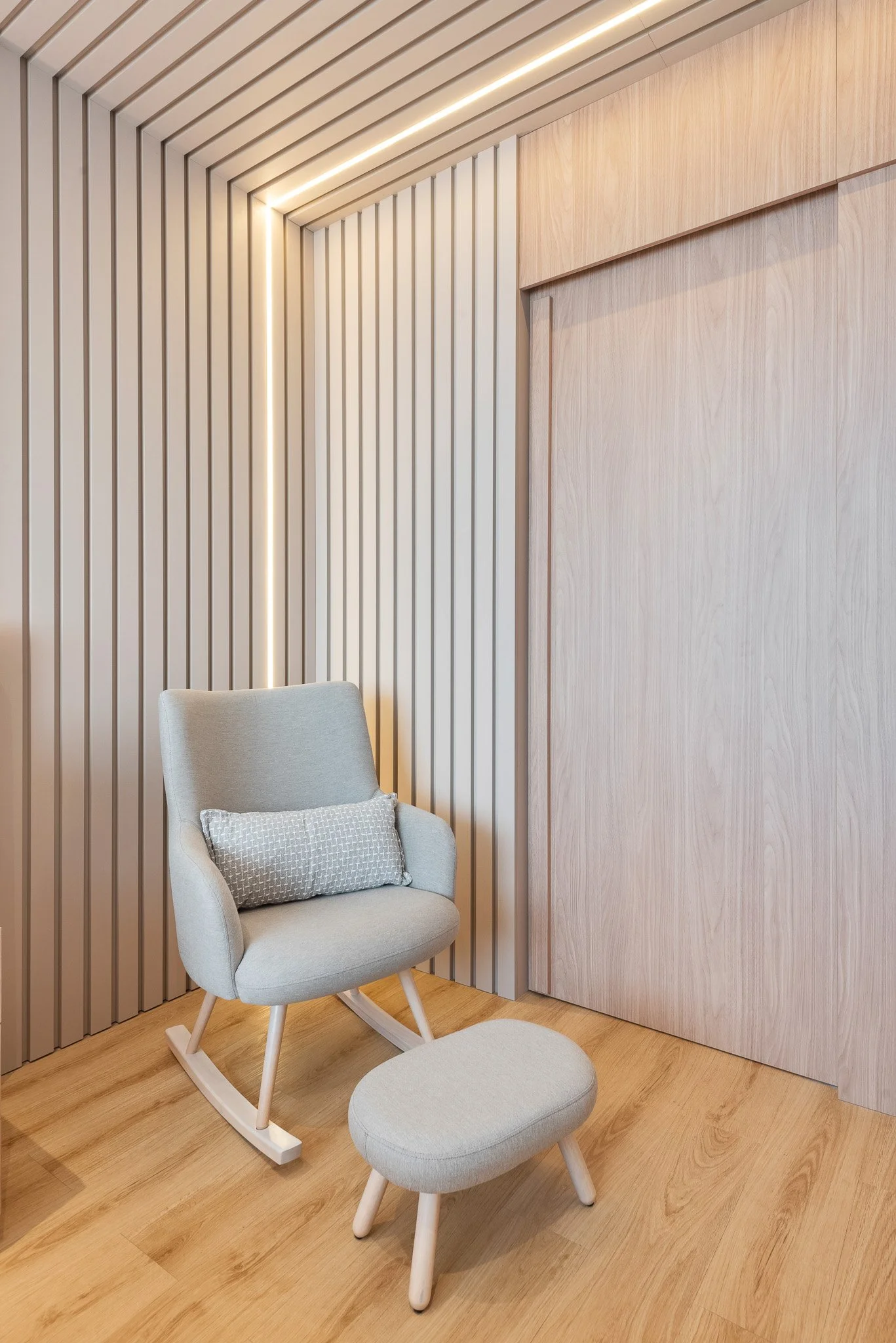 A modern beige rocking chair with a matching footrest and a textured decorative pillow, positioned in a corner with wood-paneled wall and ceiling features, and soft lighting.