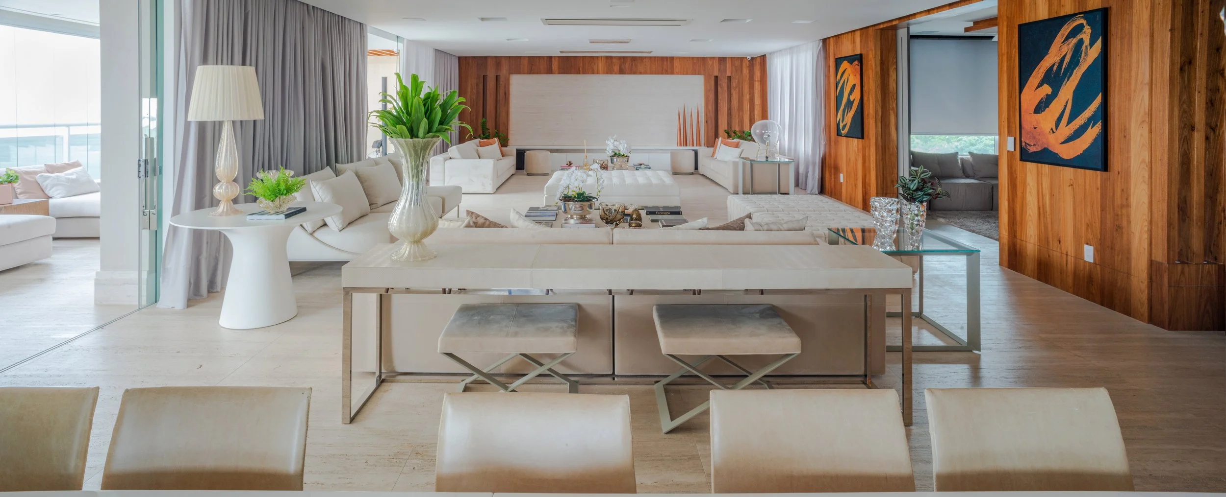 Spacious modern living room with white sofas, a large central seating area, wooden wall accents, and decorative art, with a view of the outdoors through large windows.