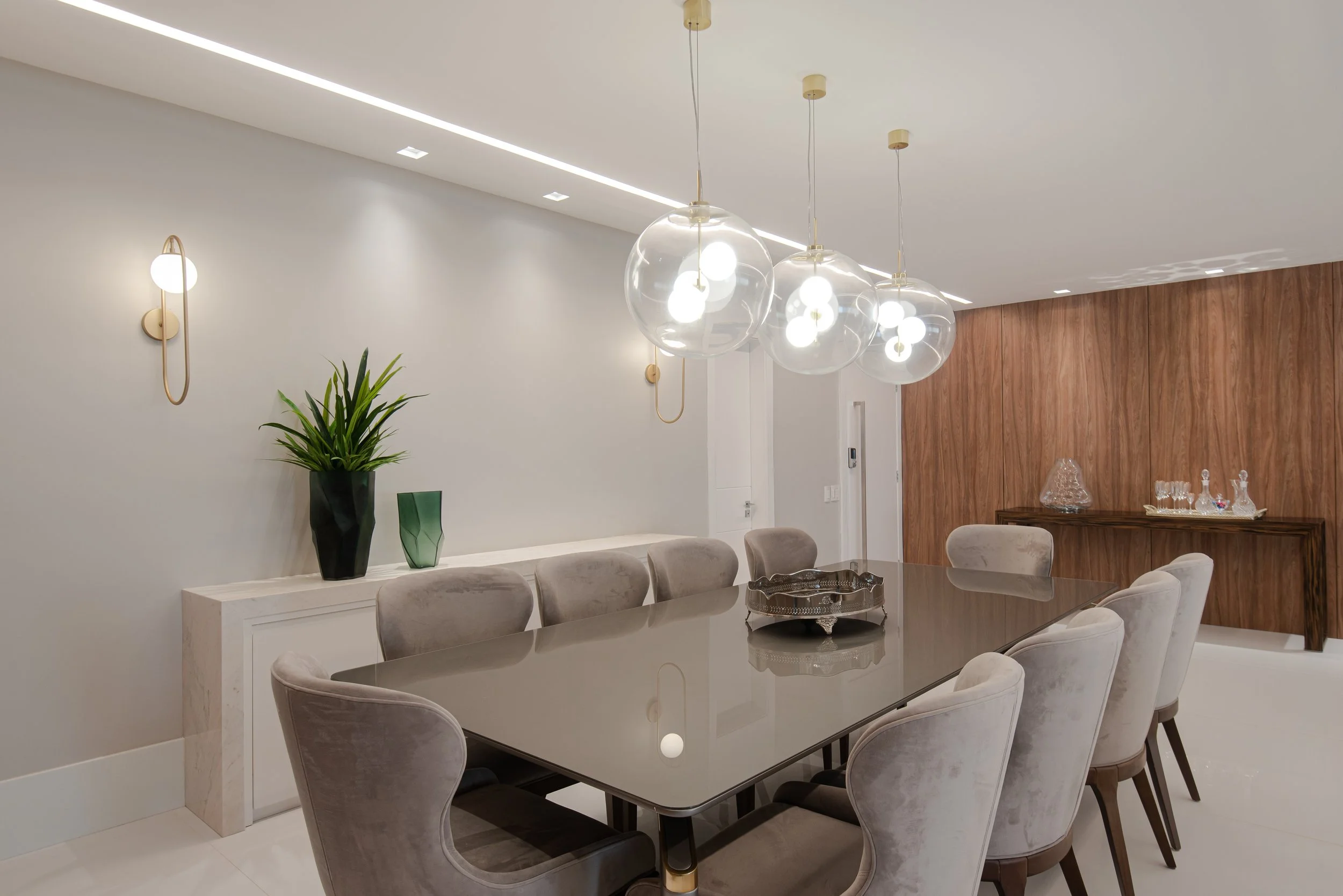 Modern dining room with a long glossy table, beige upholstered chairs, three glass globe pendant lights, a wooden accent wall, and decorative glassware on a console table.