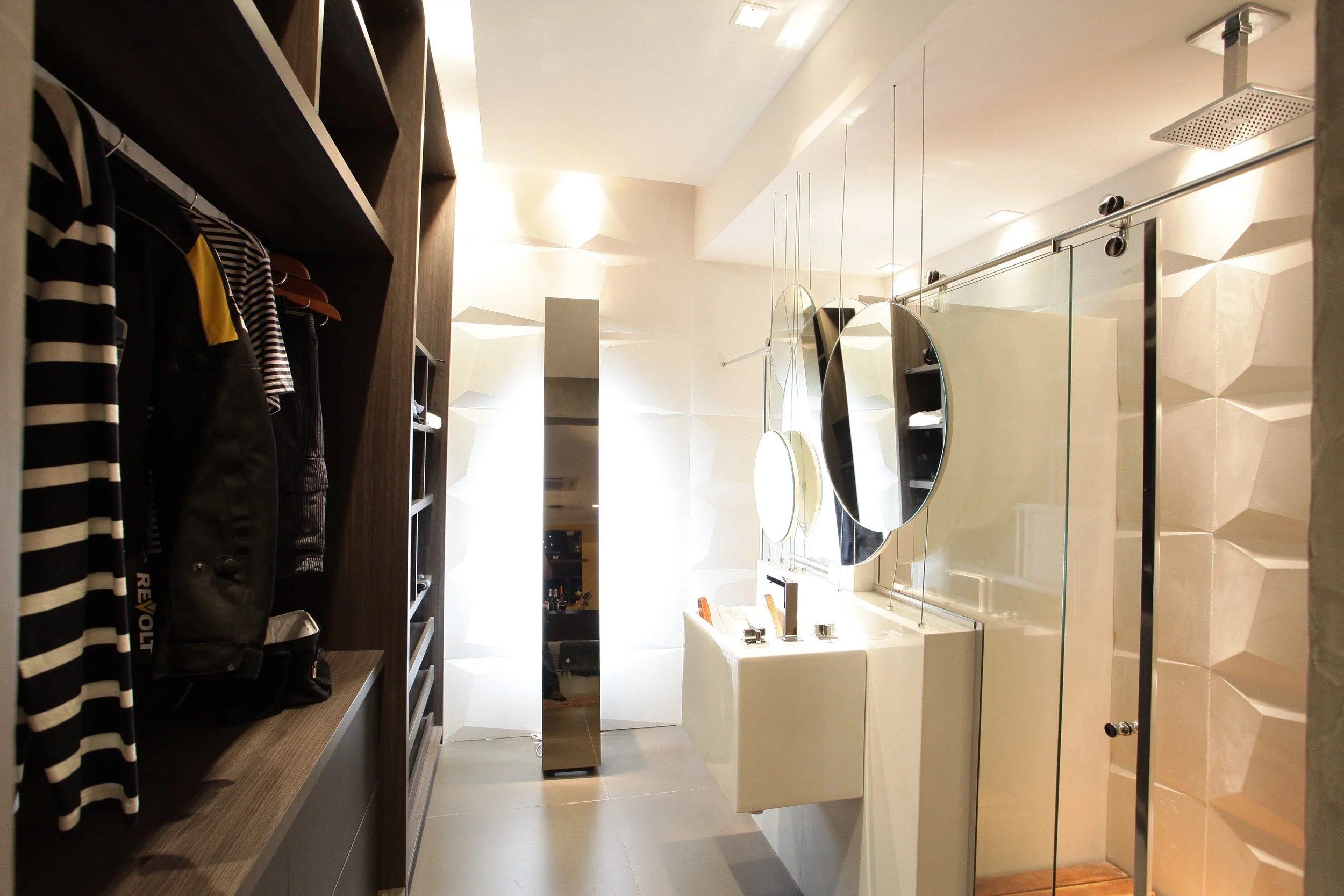 A modern bathroom with circular mirrors, a walk-in shower with a glass door, a white sink, and a textured beige wall. On the left, there is a wooden shelving unit with clothes and bags, and the floor is tiled in gray.
