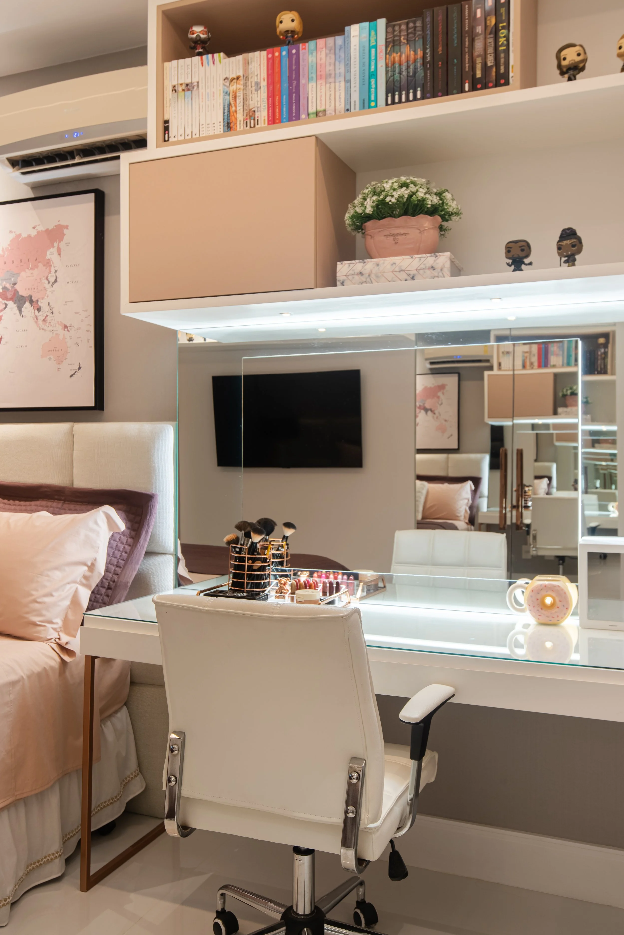A modern bedroom or dressing area with a white makeup vanity table, a white chair, a mounted flat-screen TV, and shelves with books and decorative figurines. A bed with pink and purple pillows is partially visible on the left side, and a large mirror