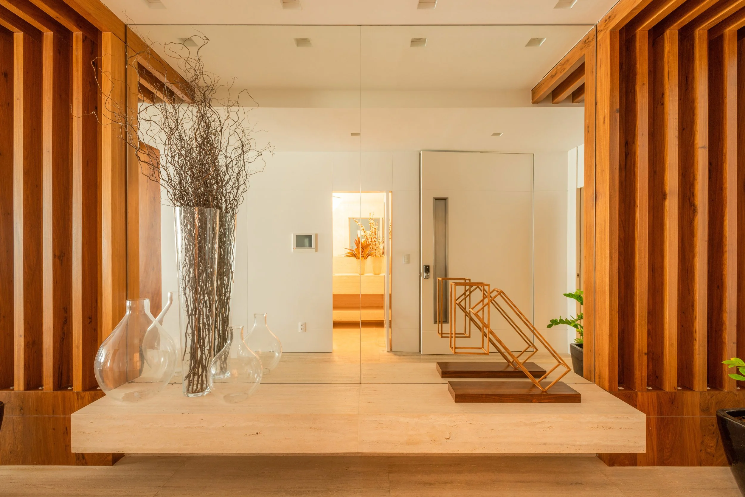 Modern entryway with wooden wall panels, a large mirror, decorative vases with branches on a beige marble table, and minimalist orange geometric sculptures, leading to a bright room with stairs and a potted plant.