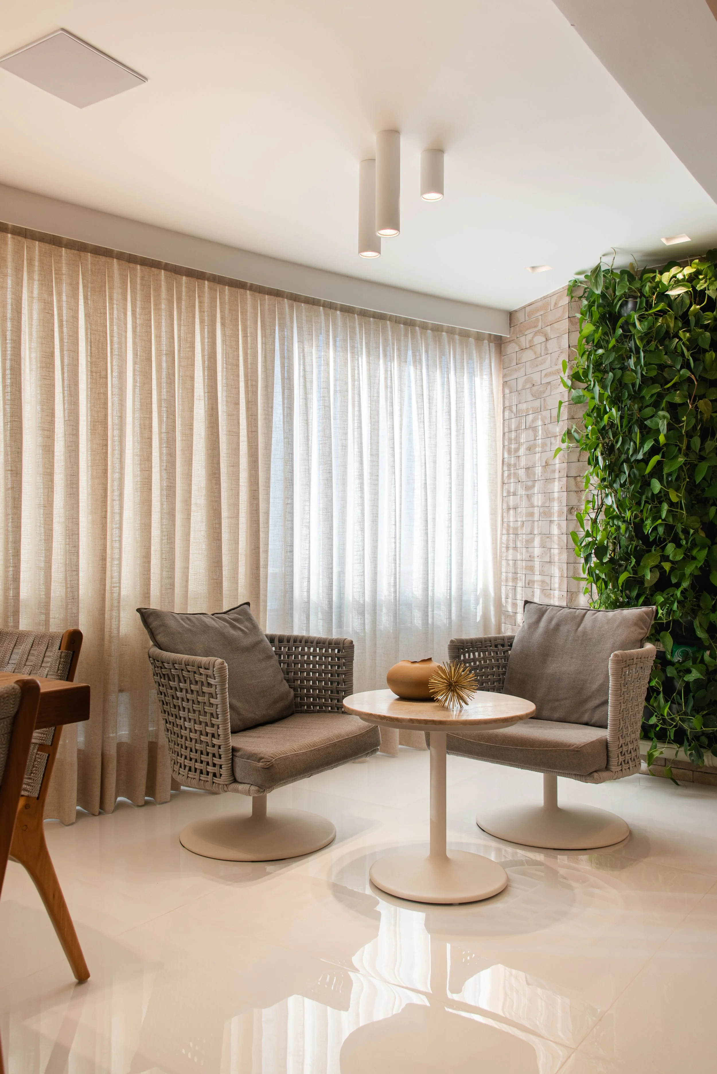 A cozy indoor seating area with two wicker armchairs with lumbar pillows, situated around a small round white table. The area features beige curtains in front of large windows, a green leafy plant on the right, and a brick wall behind the plant. The 
