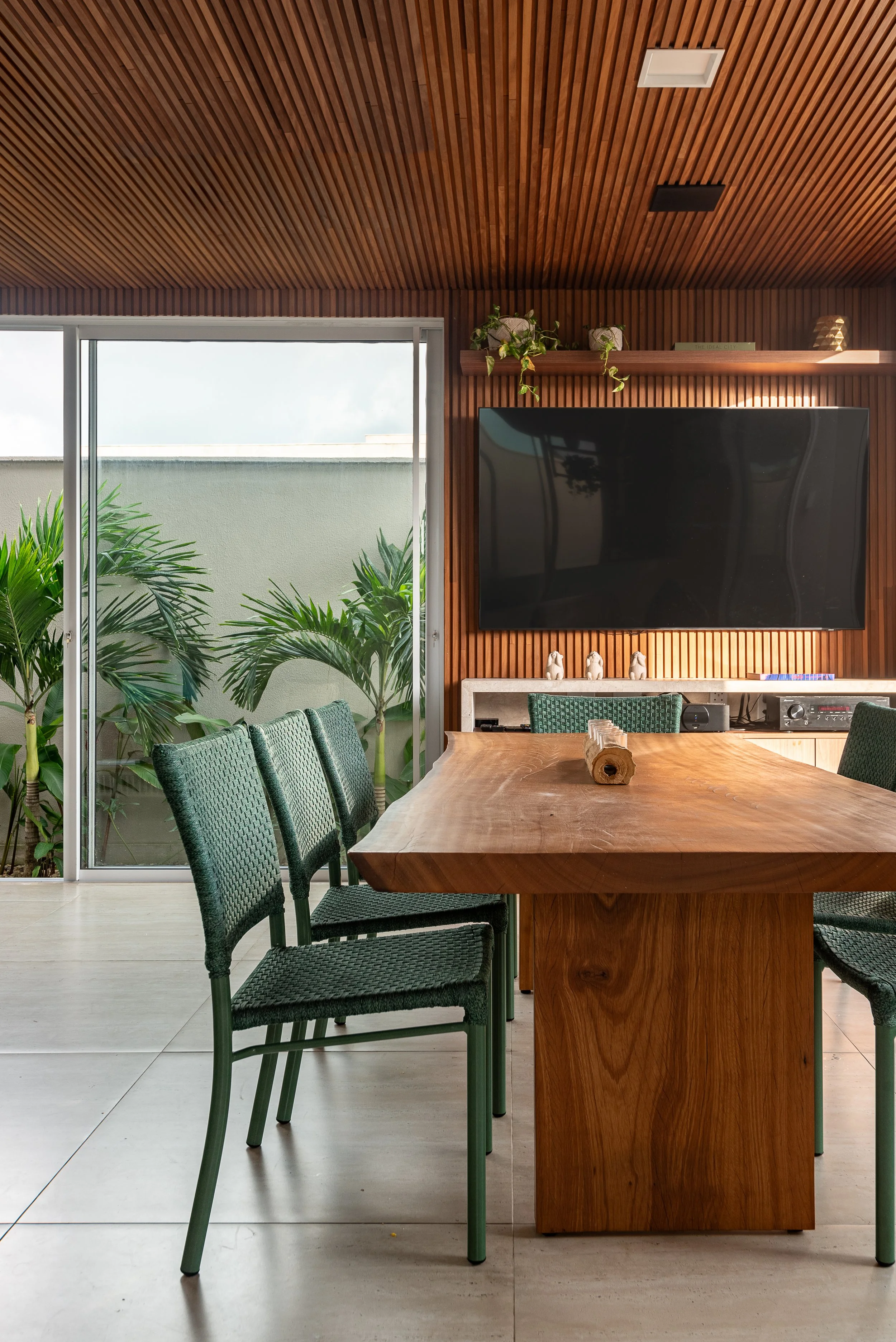 Modern dining room with a wooden table, green chairs, a large TV on a wooden wall, and sliding glass doors opening to a patio with lush green plants.