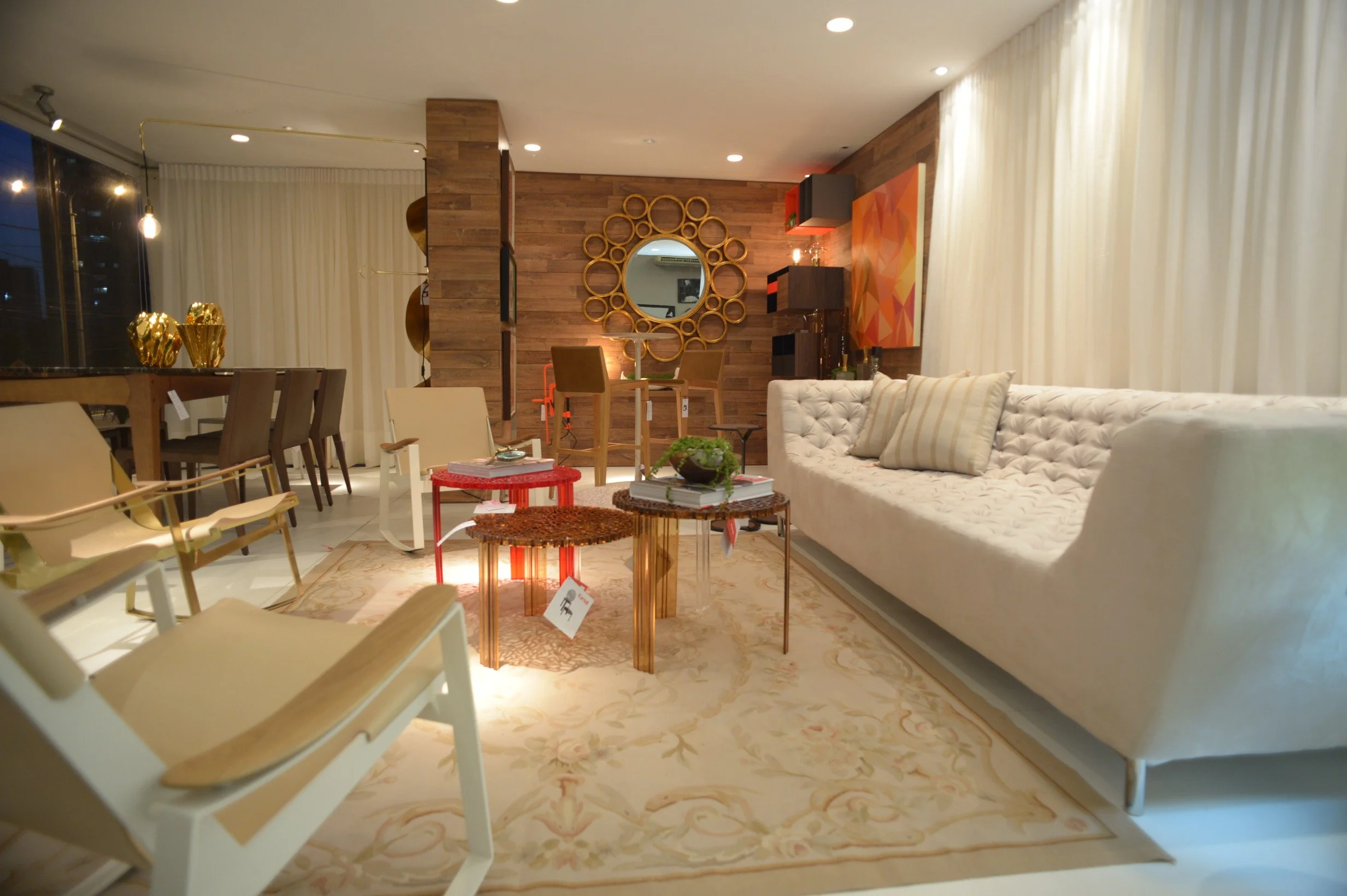 Living room with white sofa, beige and striped pillows, glass coffee table with plant, various chairs, wooden wall with circular mirror, artwork, and curtains.