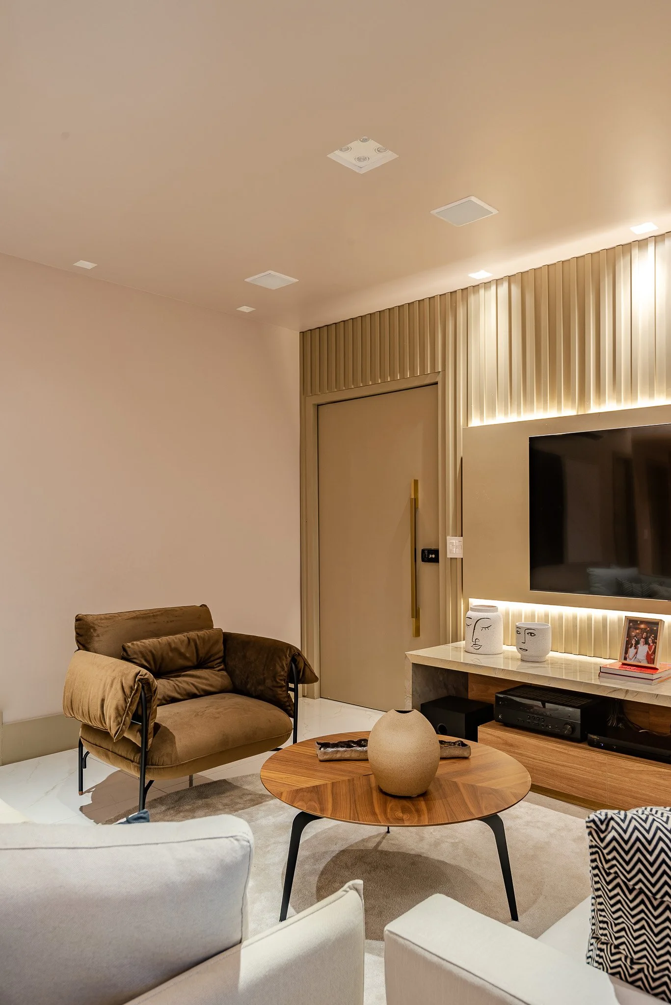 Modern living room with beige walls and ceiling, brown armchair, round wooden coffee table with a beige vase, white sofa with patterned pillow, wall-mounted TV with decorative white face vases, photo frame, and electronic devices.