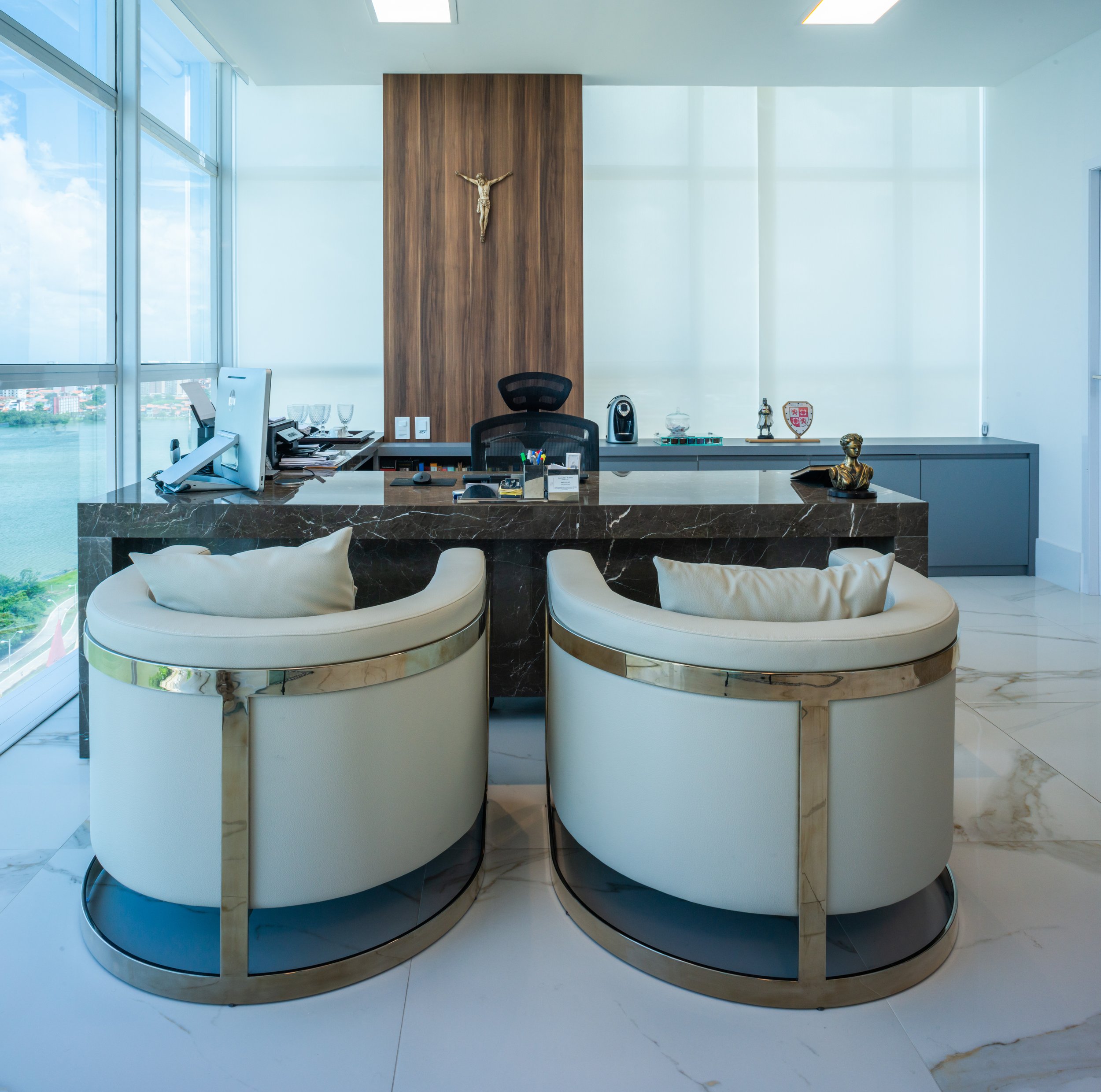 Modern office with a wooden accent wall, a crucifix, a marble desk, two white curved chairs with gold trim, a black office chair, and large windows with a view of water and cityscape.