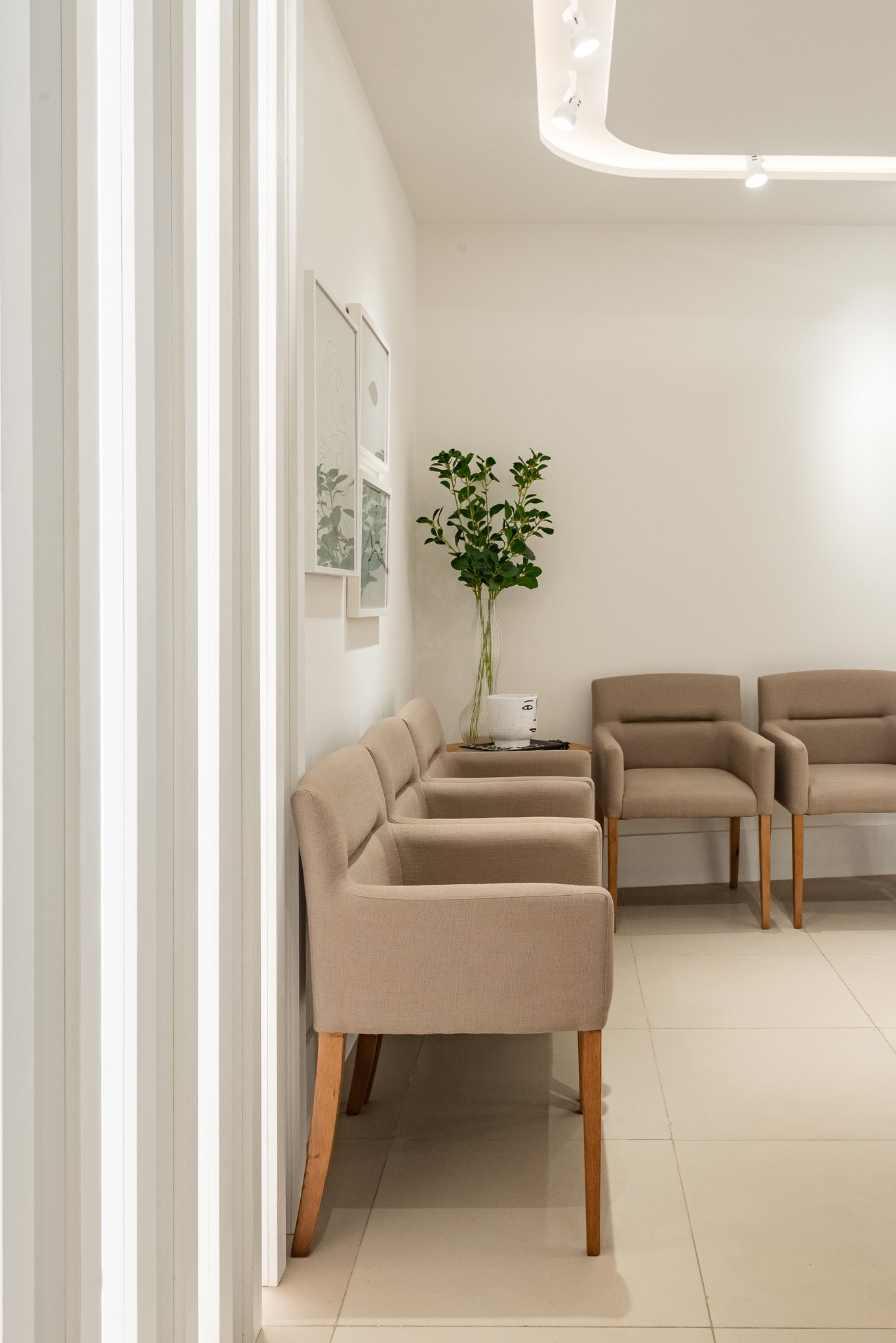 A waiting room with beige chairs, a small table with a plant, wall art, and a modern light fixture on the ceiling.