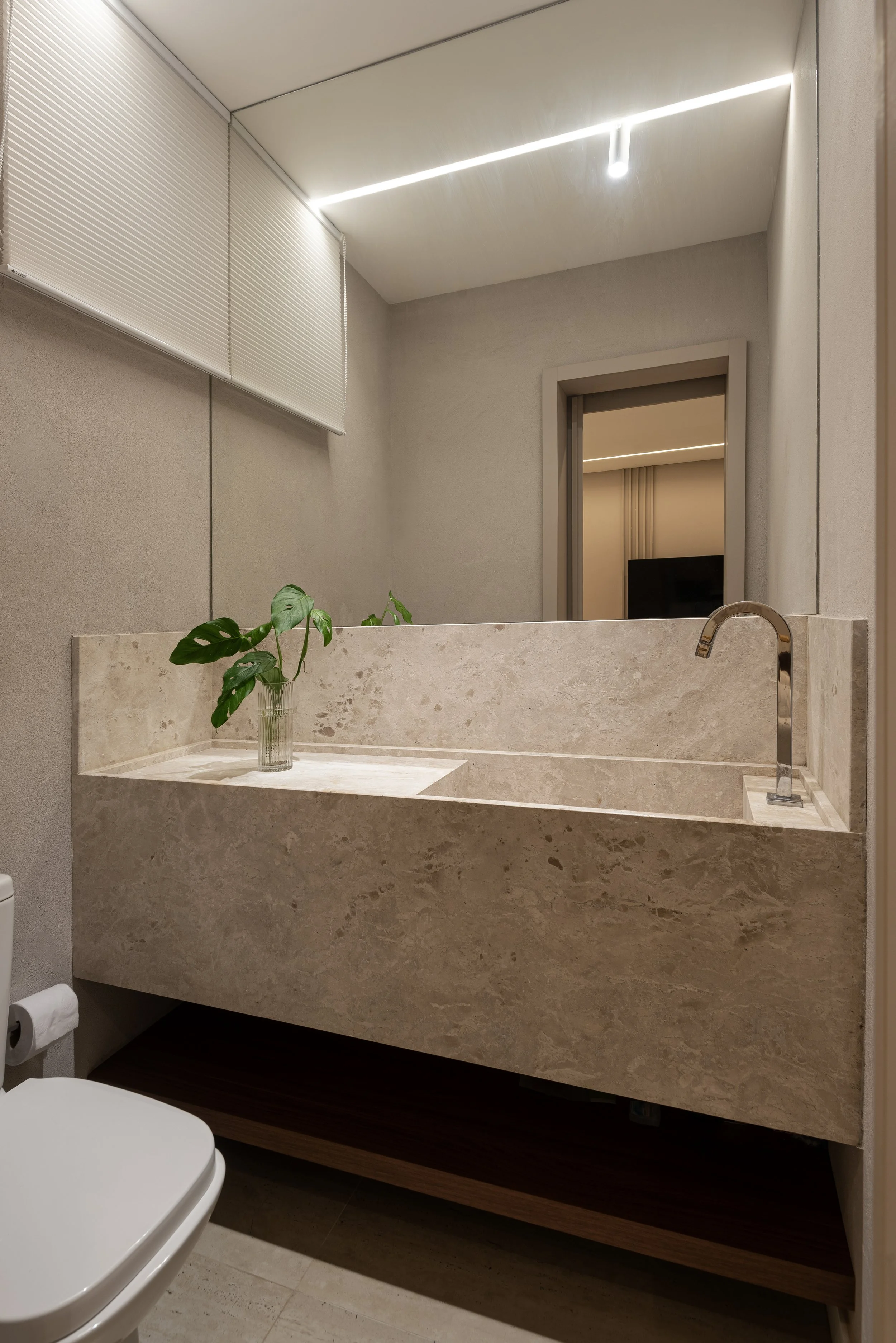 Modern bathroom with beige marble sink, a plant in a glass vase, a mirror, ceiling LED lighting, window with blinds, and part of a toilet.