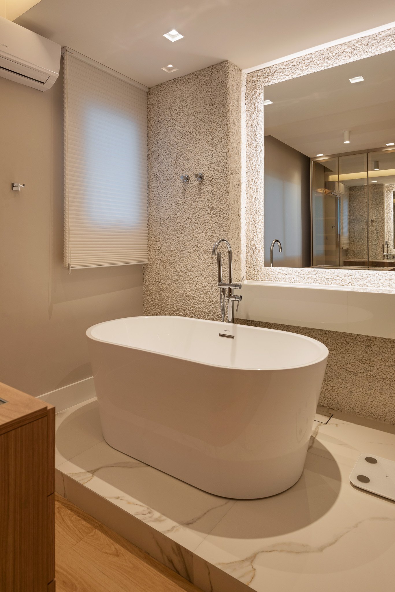 Modern bathroom with a freestanding white bathtub on a marble platform, a large mirror, textured wall, window with blinds, and a digital scale on the marble floor.