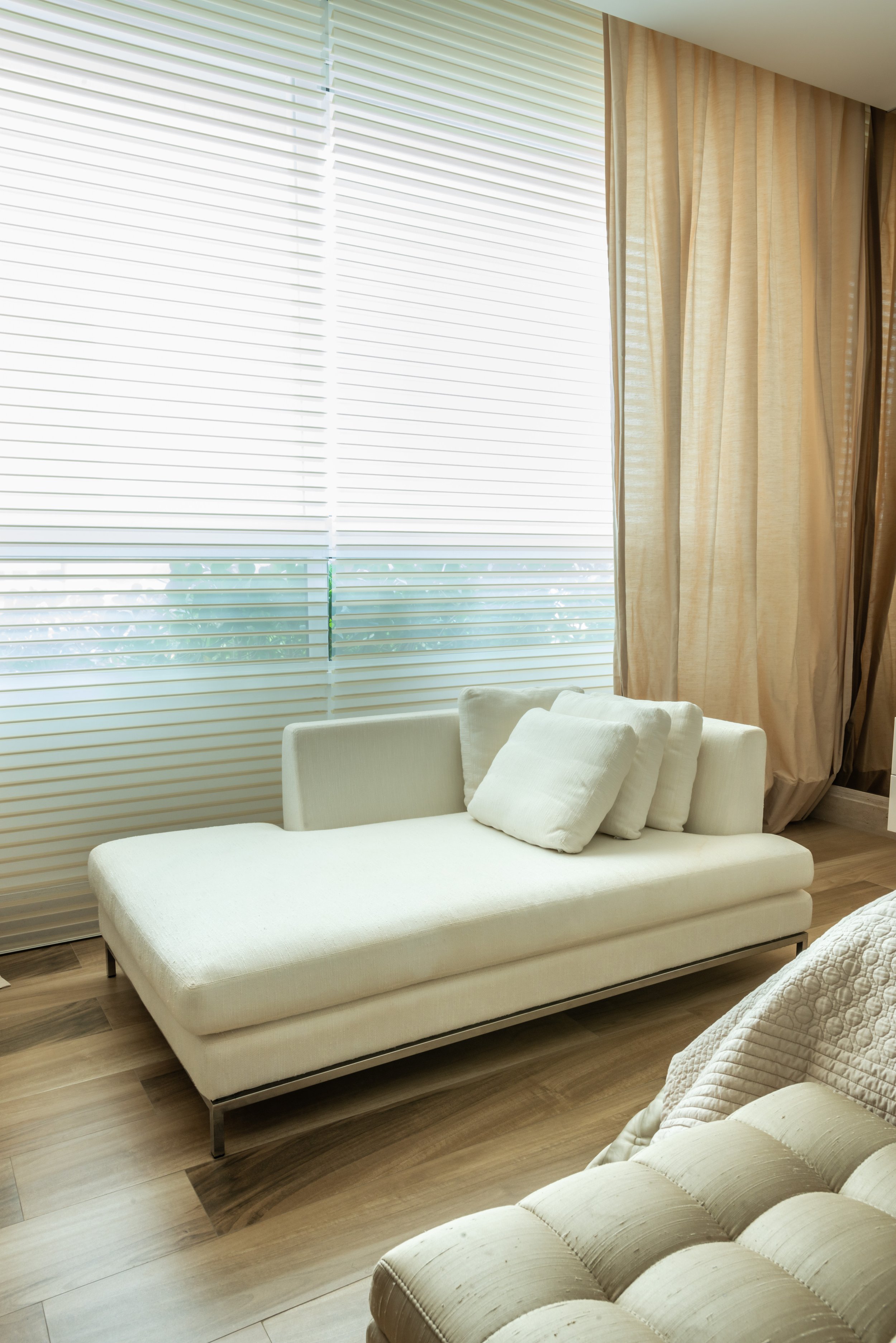 A bright, modern living room with a white chaise lounge and several white pillows, large windows with white blinds, beige curtains, and wood flooring.
