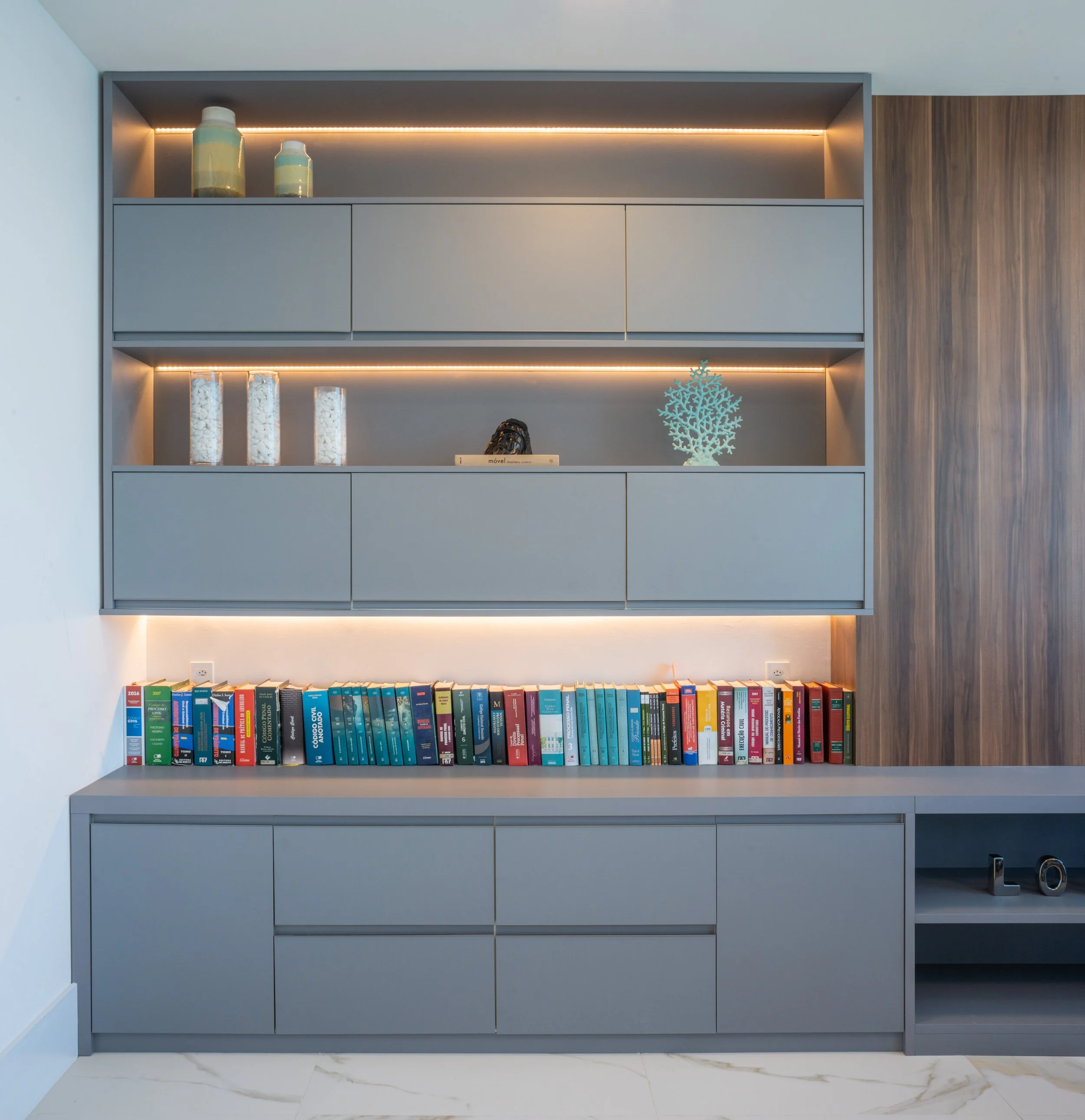 Modern gray cabinet with books, decorative items, and built-in lighting, against a wood-paneled wall in a living room.