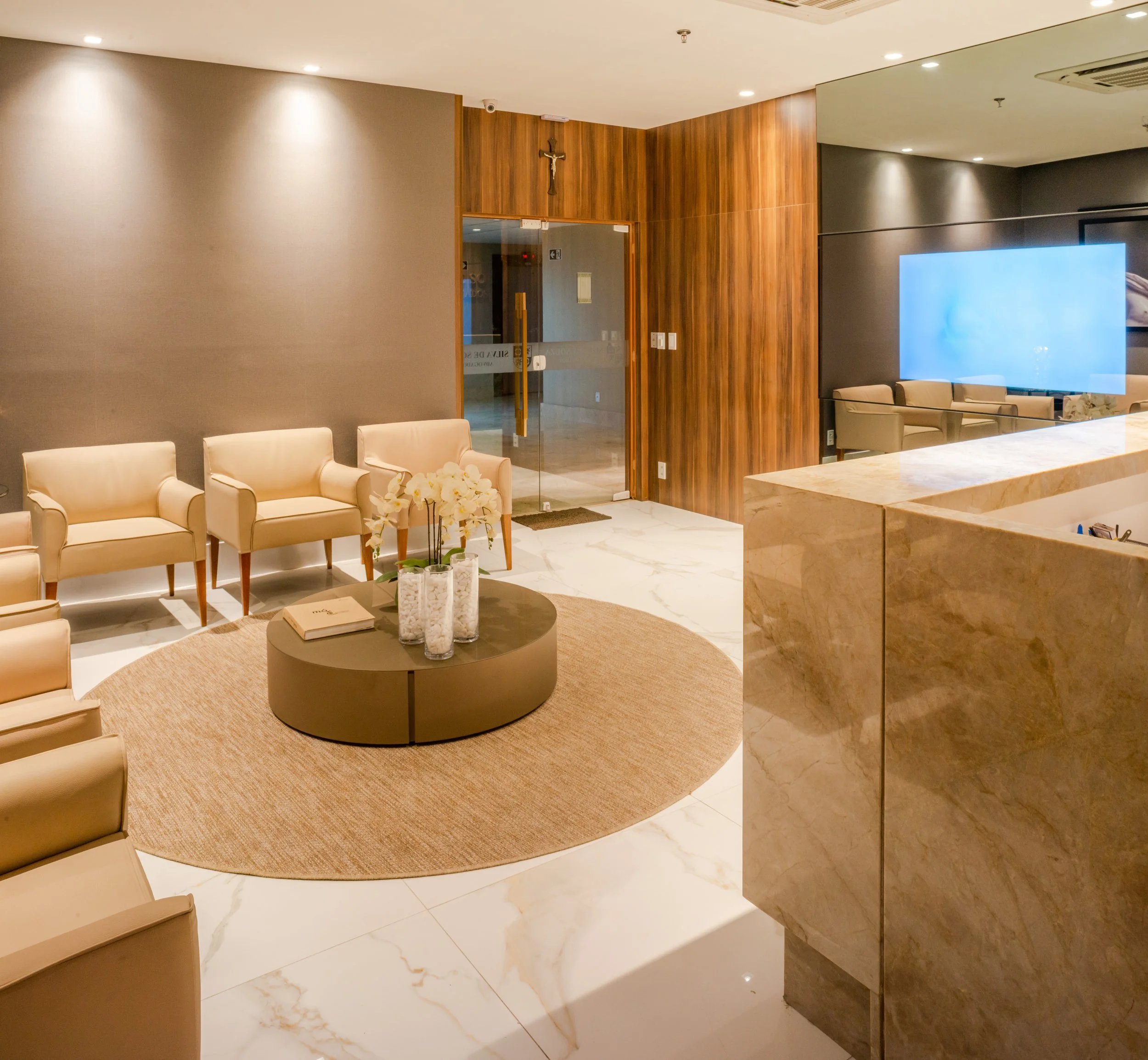 A modern hotel lobby with beige chairs, a round coffee table with flower vases, a beige rug, marble flooring, a wooden wall with a cross, glass doors, and a large screen on the wall.