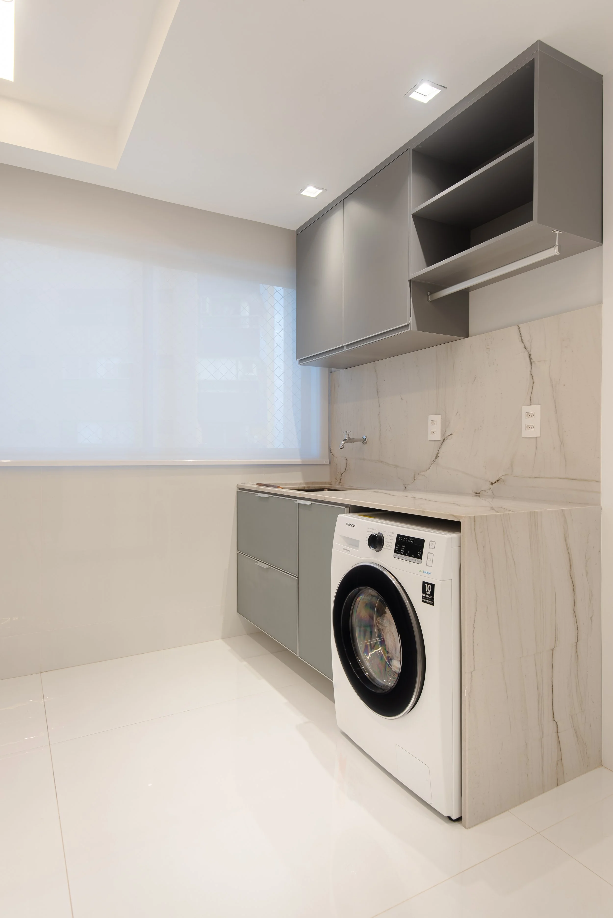 Modern laundry room with white tile floors, marble backsplash, gray cabinets, washing machine, and large window with a white roller shade.