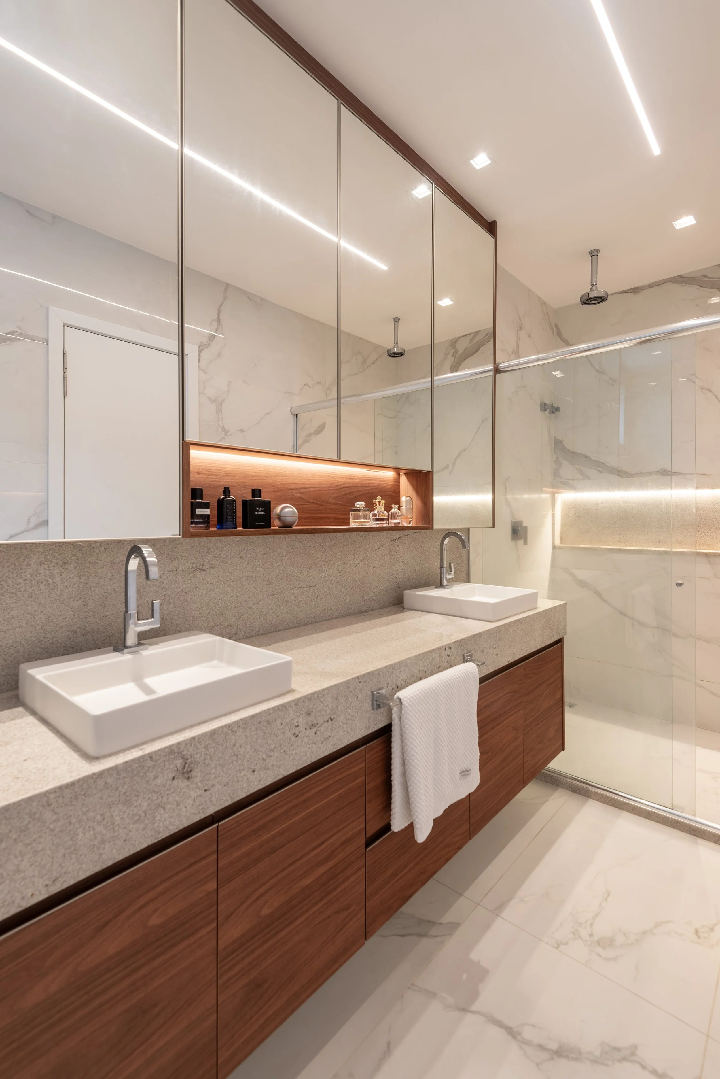 Modern bathroom with beige countertops, double sinks, wooden cabinet below, large mirrored cabinet above, marble walls, glass shower enclosure with marble interior, white towel hanging on cabinet.