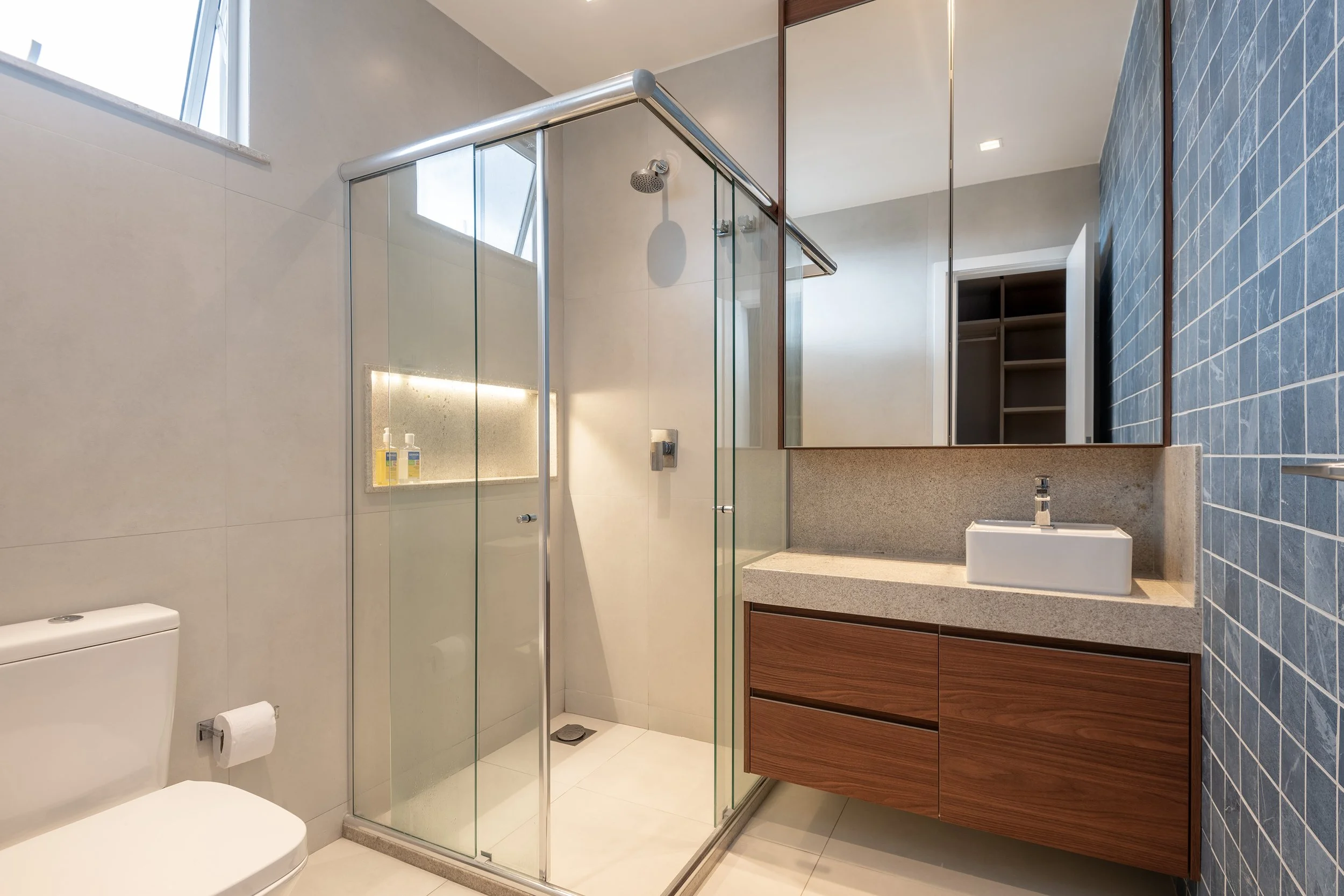 Modern bathroom with a glass shower enclosure, white toilet, wooden vanity with a rectangular sink, large mirror, and blue tiled accent wall.