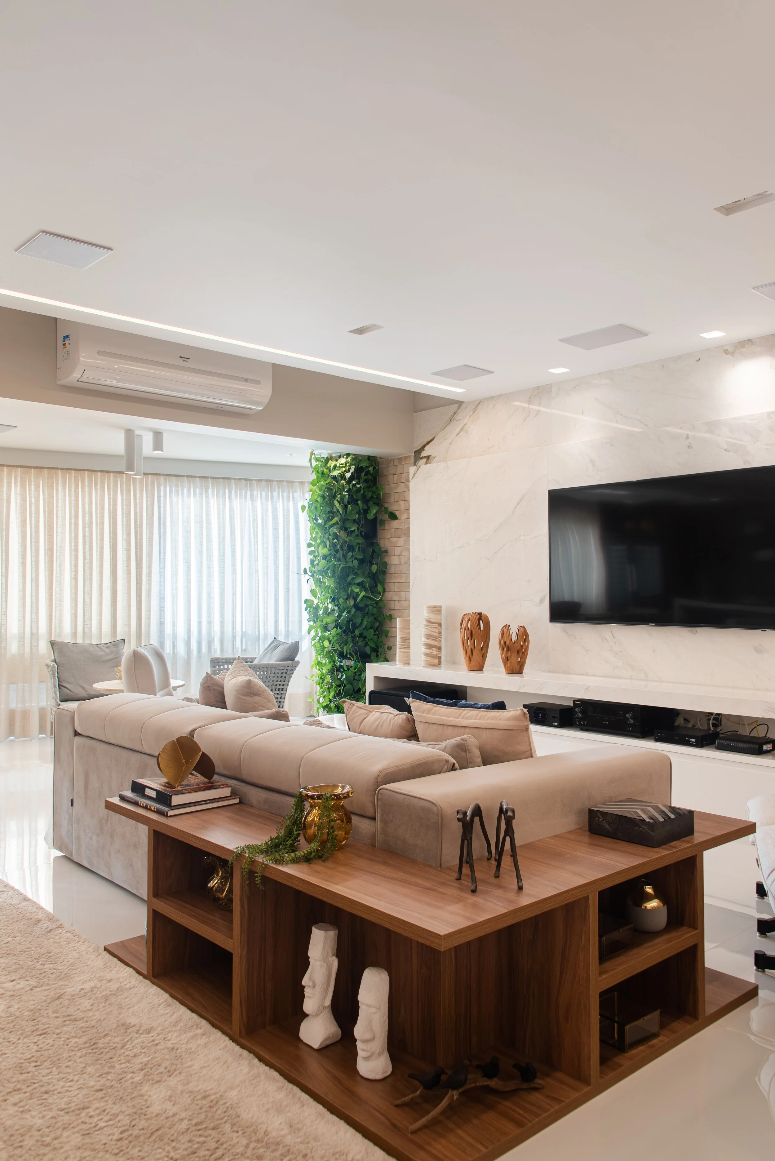 Modern living room with beige sectional sofa, wooden coffee table, decorative items, and a mounted flat-screen TV on a marble wall.