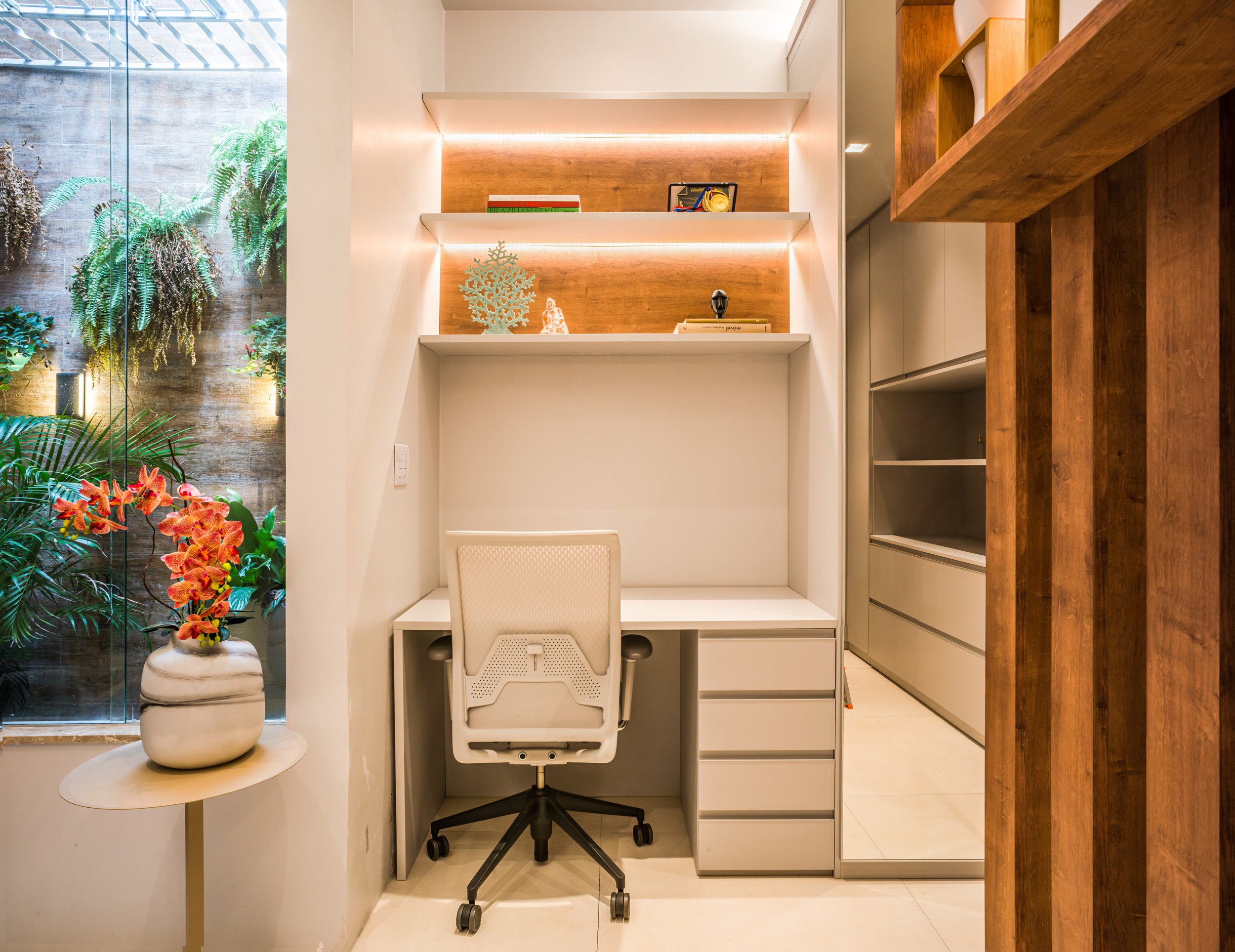 A modern home office with a white desk and an ergonomic white chair. There are built-in shelves with decorative items above the desk, and a small table with a potted plant to the left. A wood-paneled wall is to the right, and a large window with gree