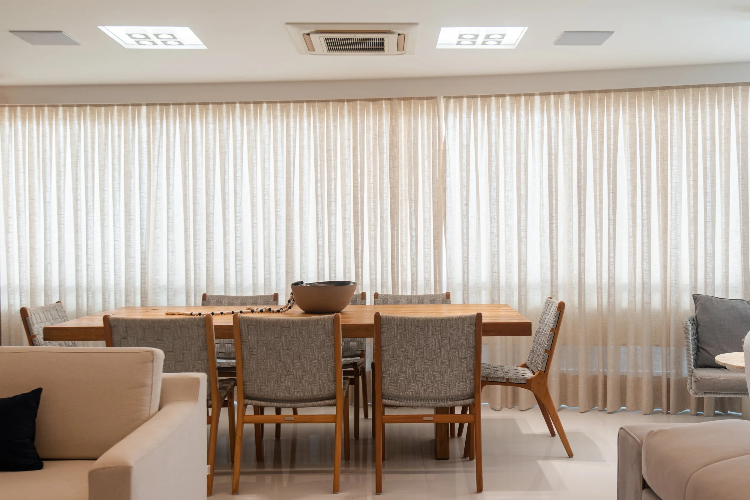 Dining area with wooden table, gray upholstered chairs, beige curtains, and light-colored sofas in the foreground.