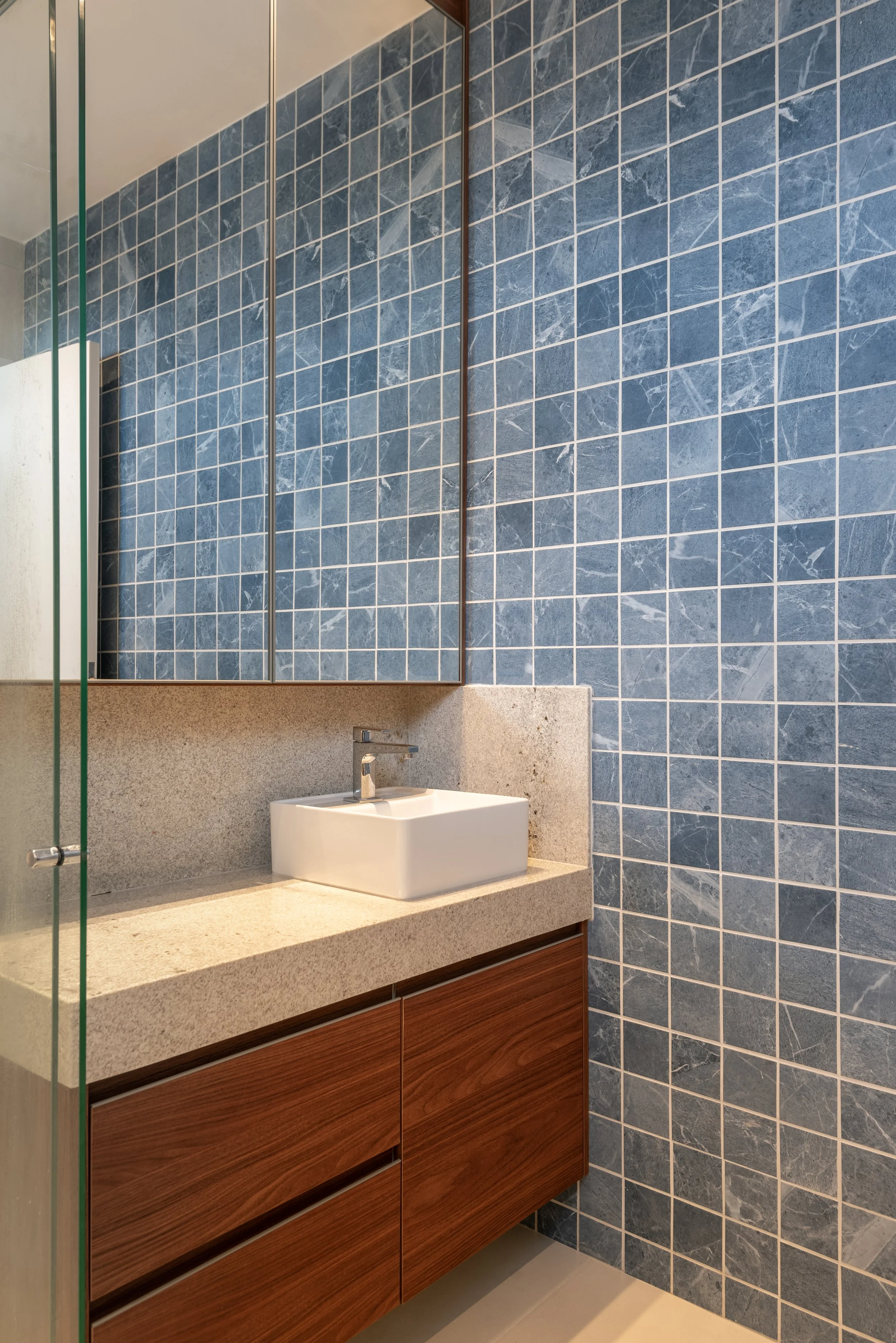 Bathroom with a blue tiled wall, a floating wooden vanity, a speckled countertop, a white square vessel sink, a chrome faucet, and mirrored cabinet doors.