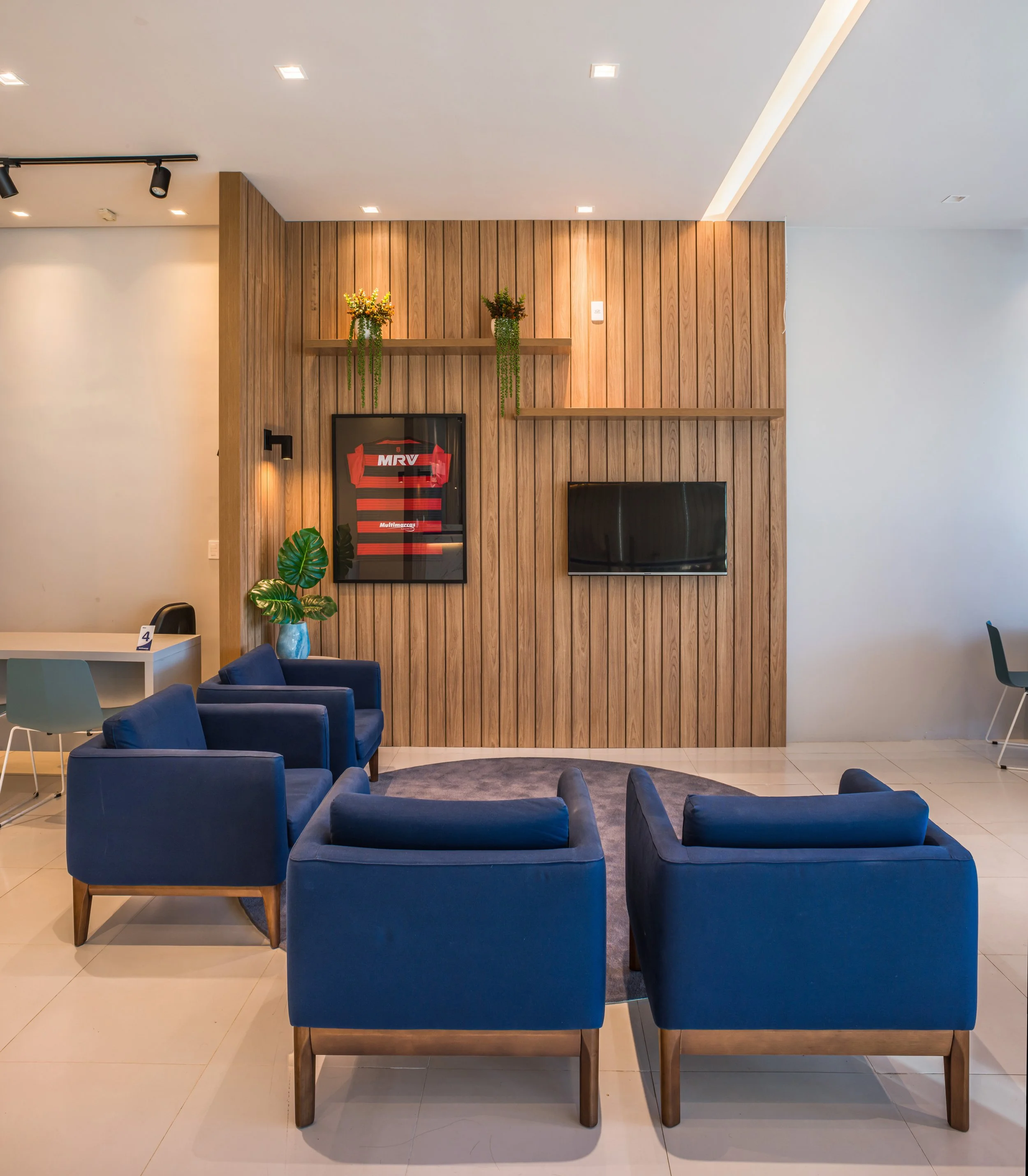 Modern living room with six blue armchairs arranged around a small circular rug in front of a wooden feature wall, with a flat screen TV, framed sports jersey, potted plant, and floating shelves with plants.