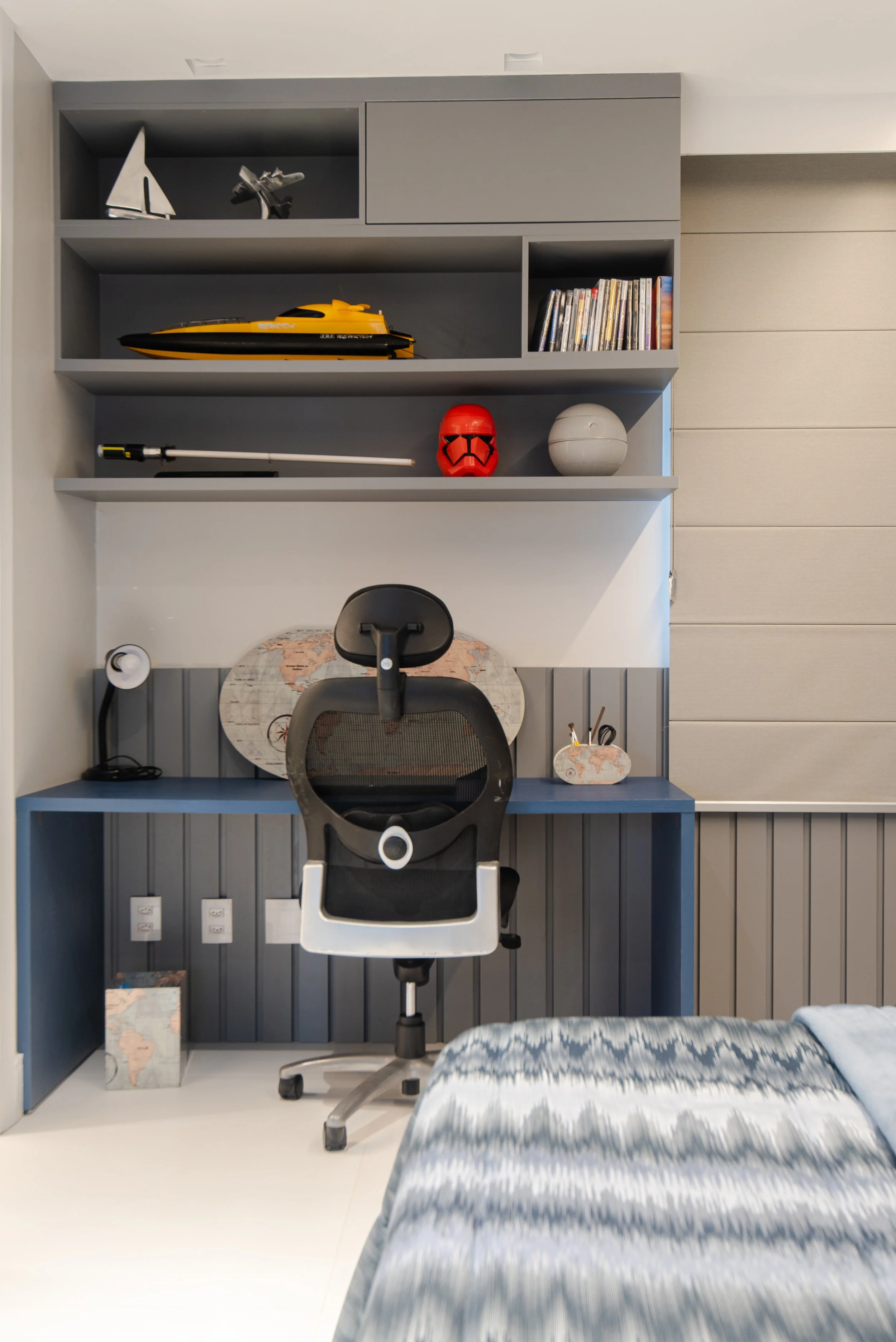 A bedroom workspace with a blue desk and ergonomic black office chair, featuring shelves above with a model sailboat, a toy yacht, a red Stormtrooper helmet, and books, with a world map in the background.