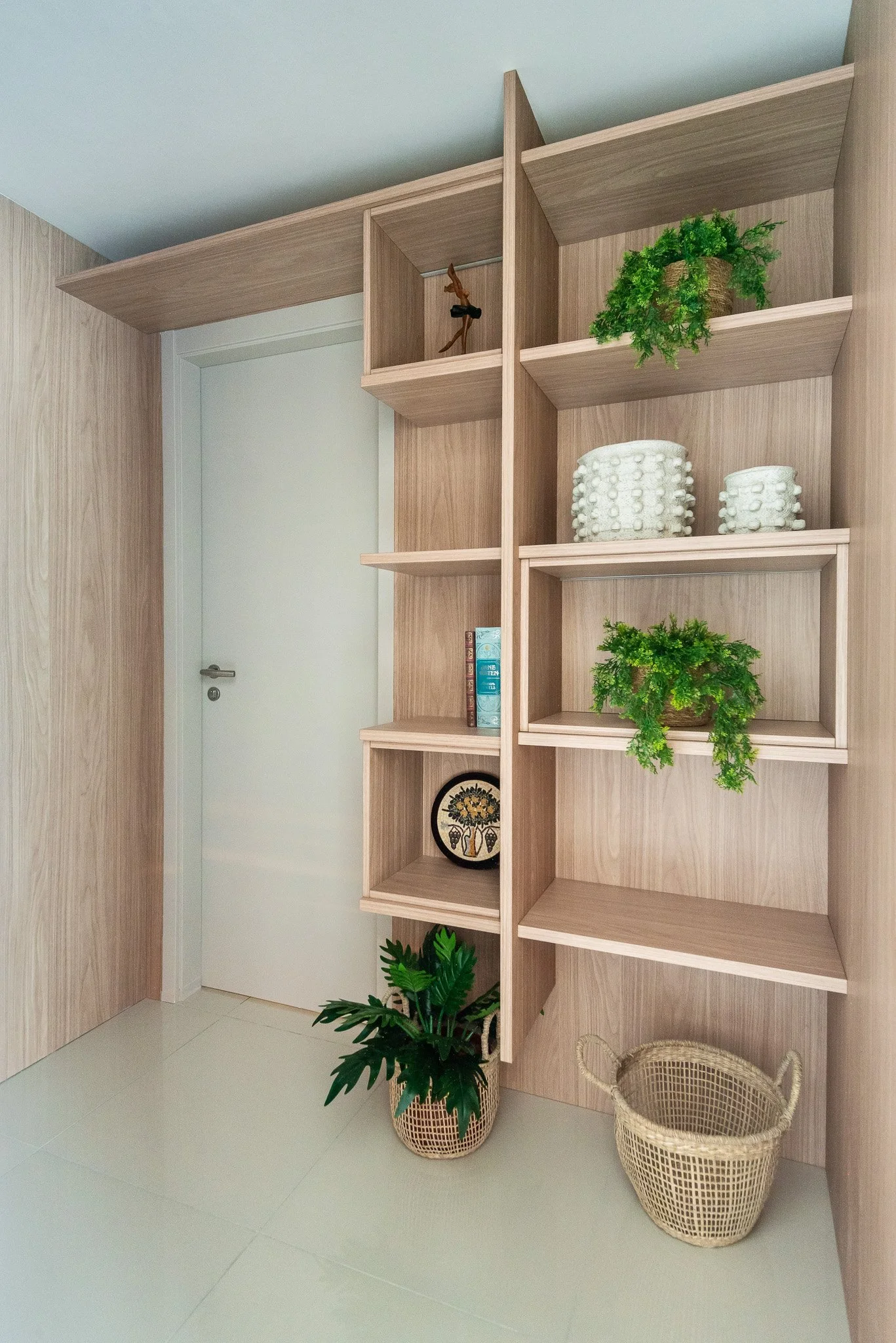 Wooden shelving unit against wall with decorative items, plants, books, and baskets, next to a white door on a tiled floor.