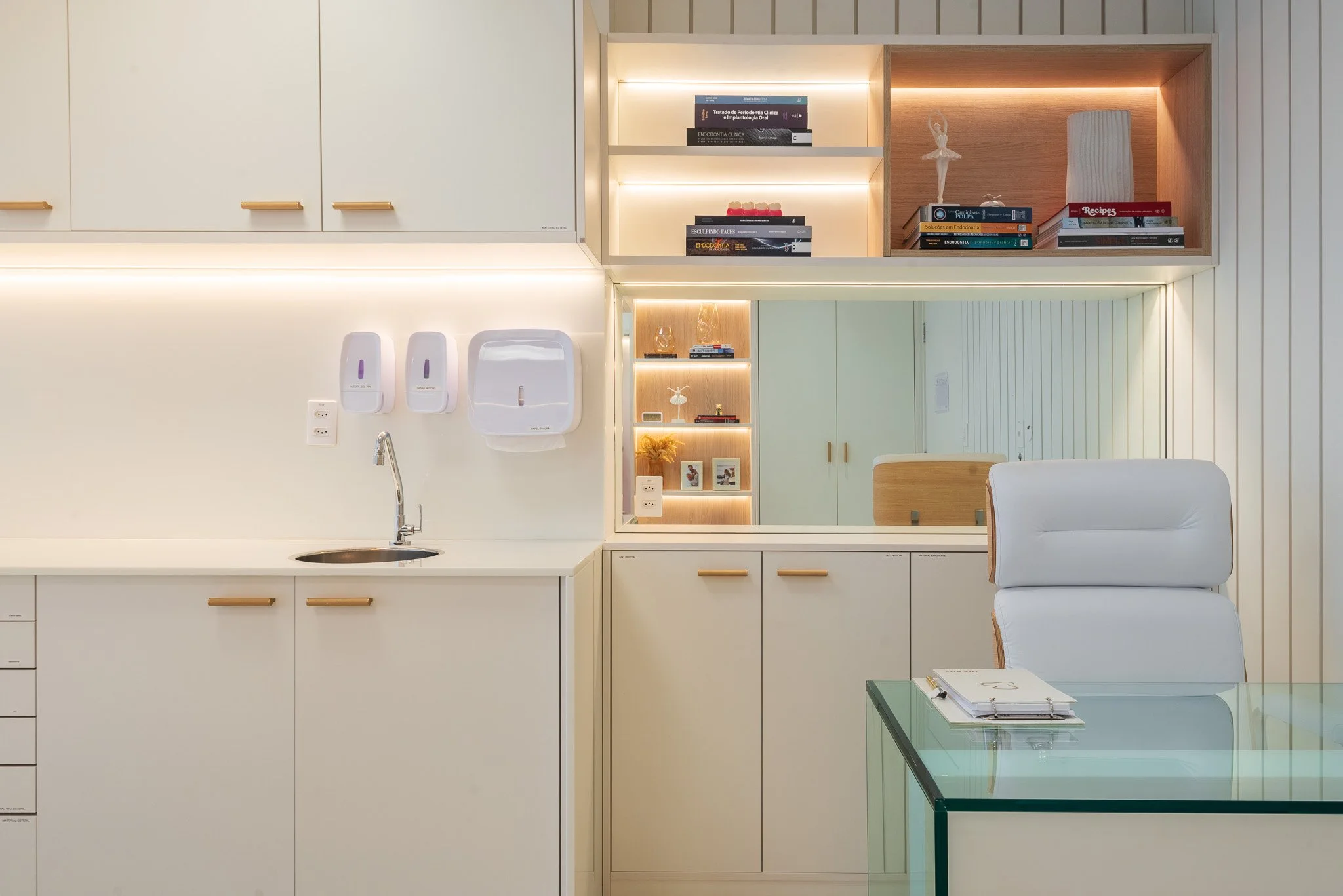 A modern office or medical reception area with white cabinetry, a glass-topped table, and shelves with books and decorative items. There is a mirror on the wall, and a white armchair is partially visible.