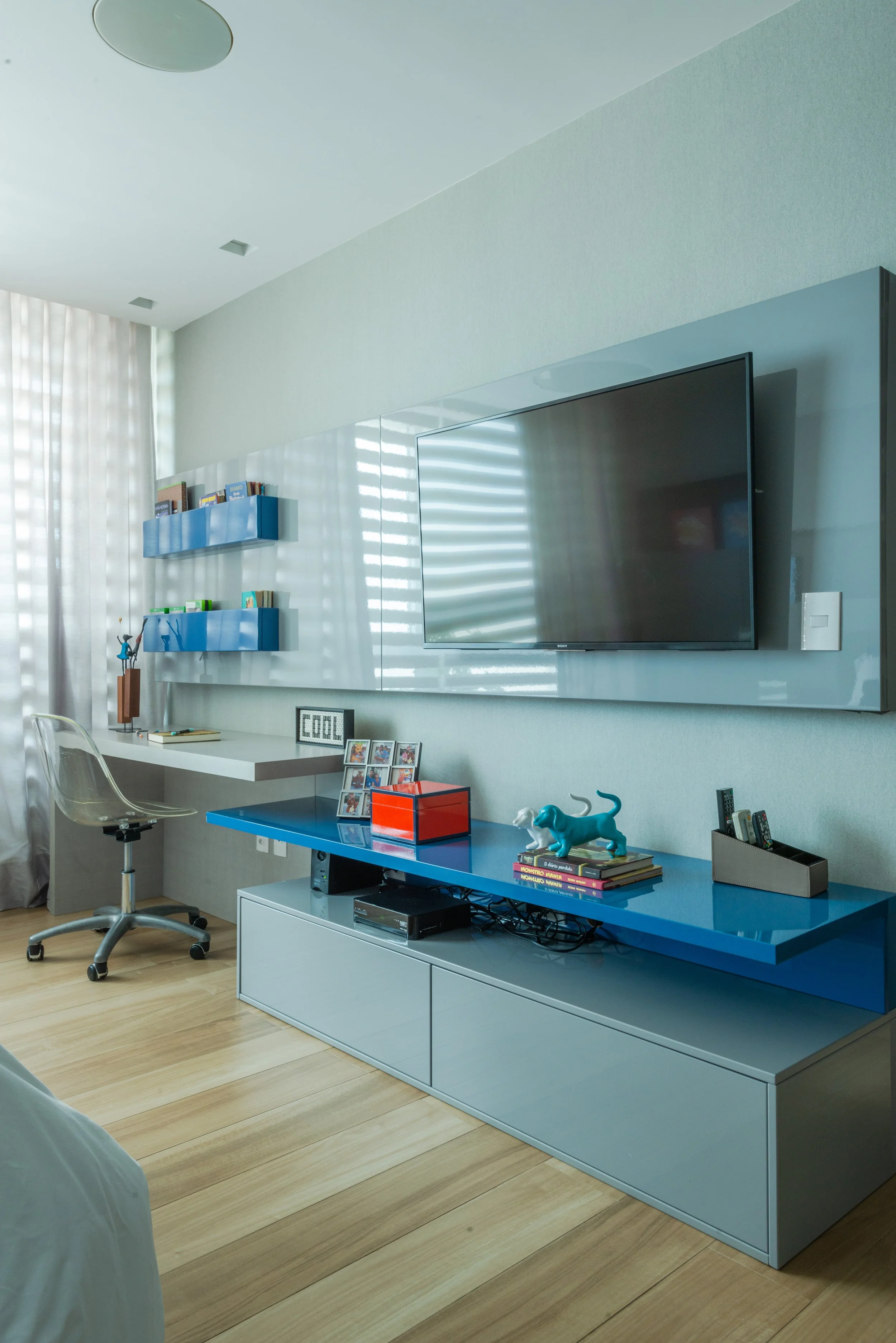 Modern living room with a wall-mounted flat-screen TV, a white and blue media console, decorative items like a blue sculpture, framed photo collage, books, and a remote holder. There is a white desk with a transparent chair, blue floating shelves wit