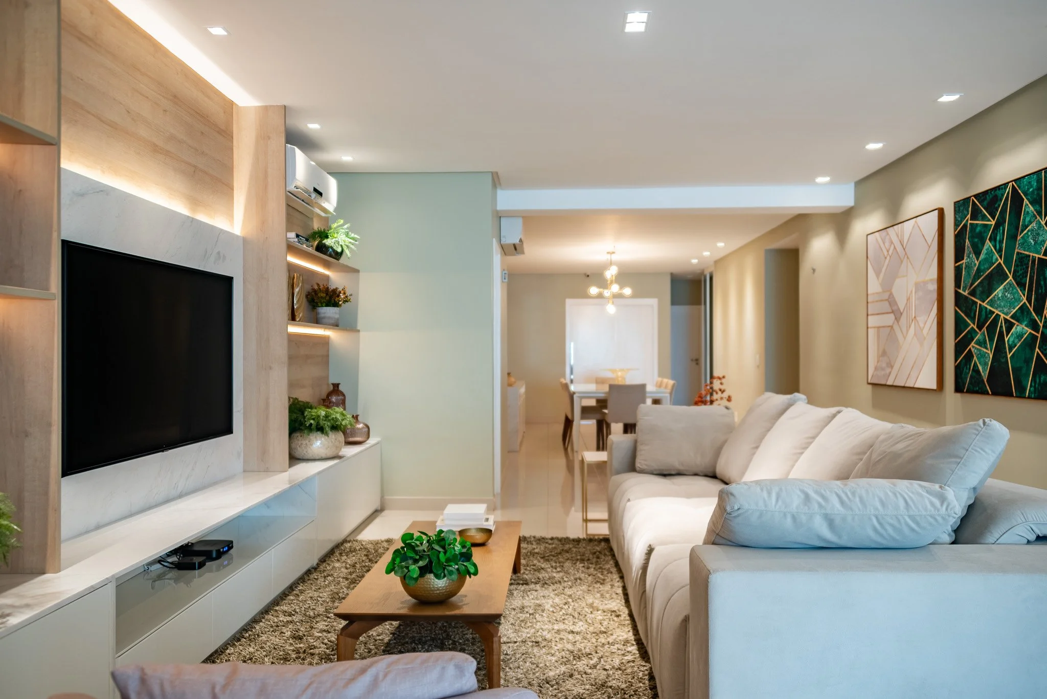 Living room with beige sofa, coffee table, wall art, and built-in entertainment center with TV, shelves, and plants, leading to dining area with chandelier.