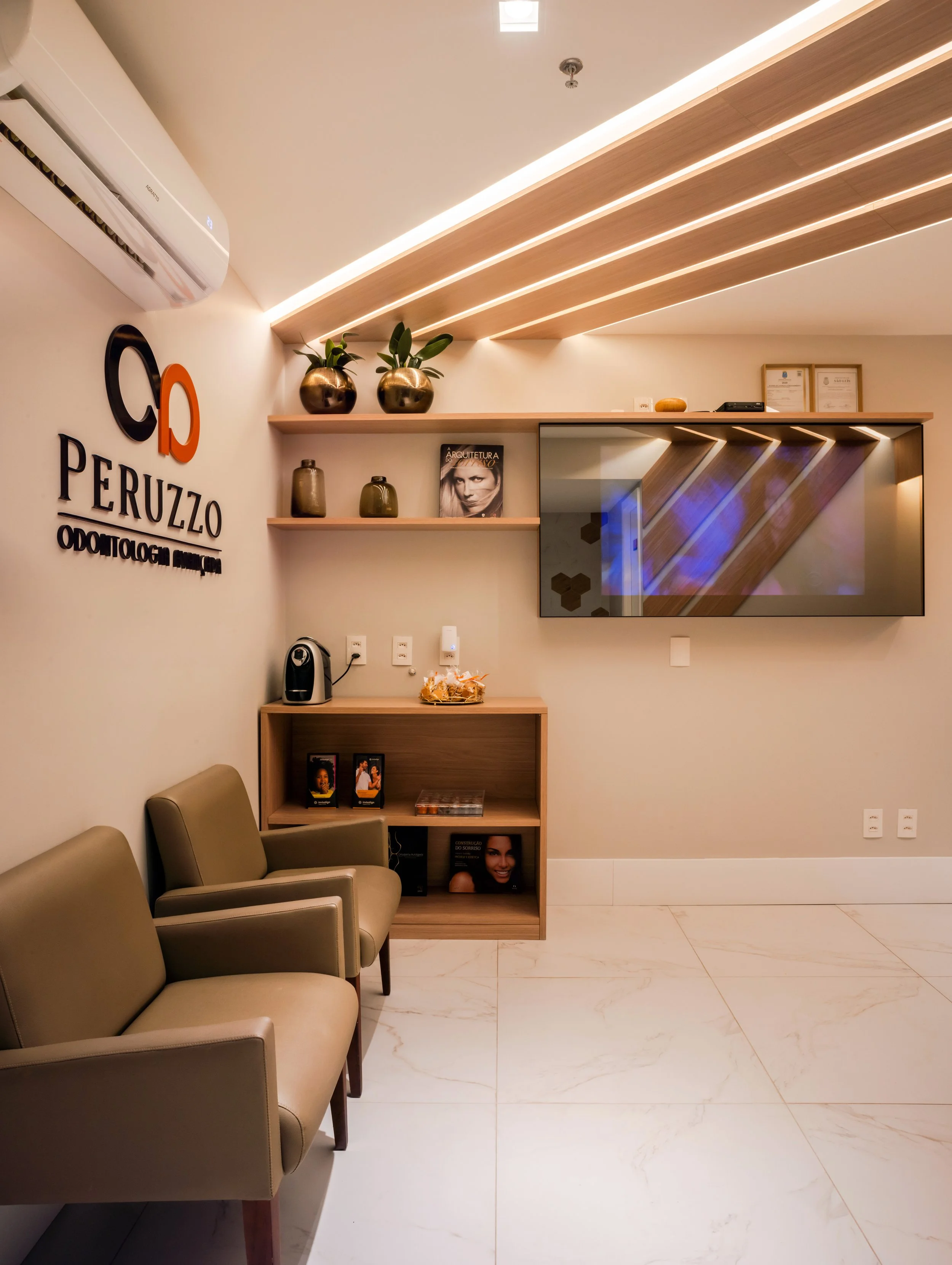 Modern waiting area with two beige chairs, shelves with decorative vases and magazines, wall-mounted TV, framed documents, and an air conditioner at the top.
