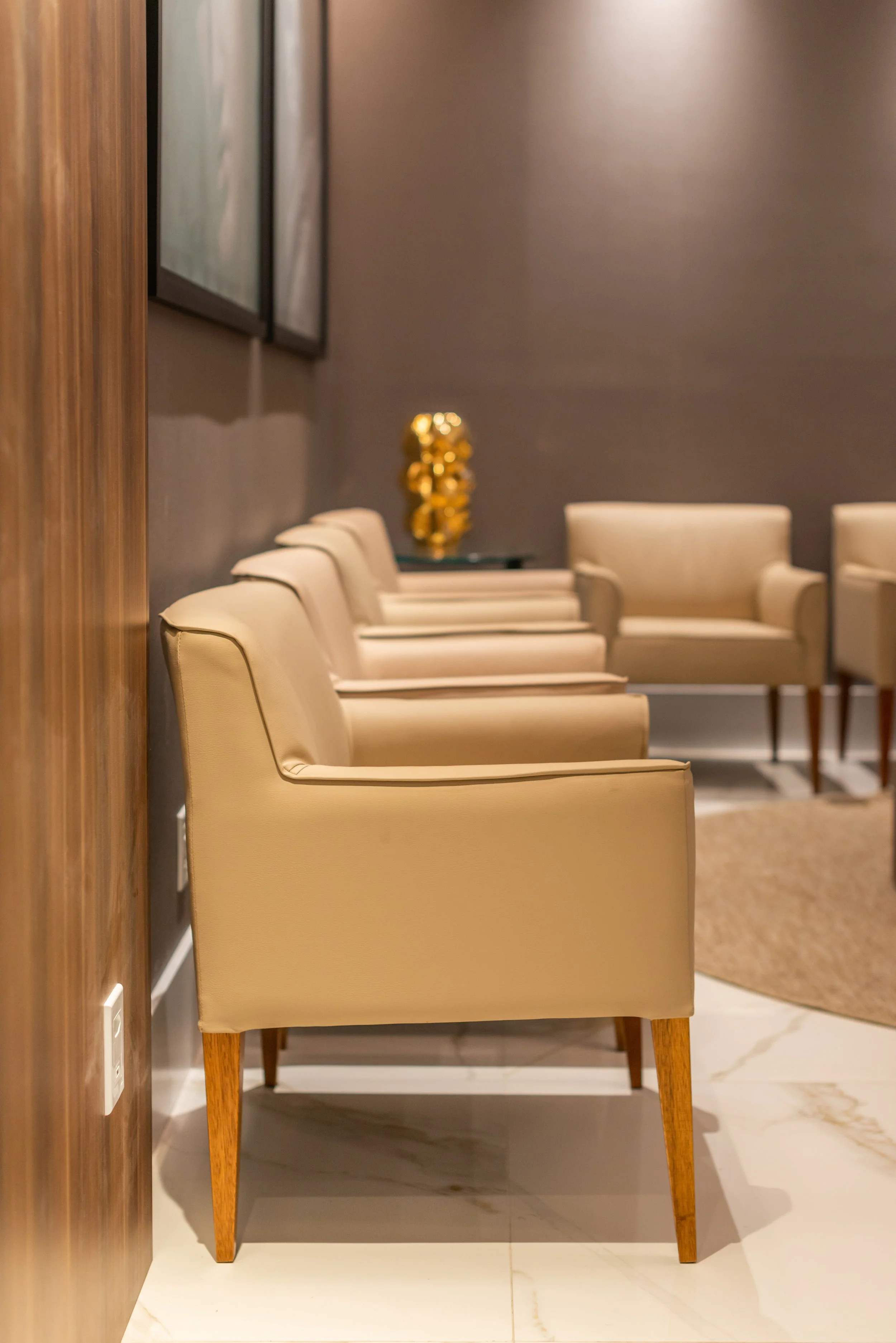 A row of beige leather armchairs in a waiting area with a dark wall and a gold decorative sculpture in the background.