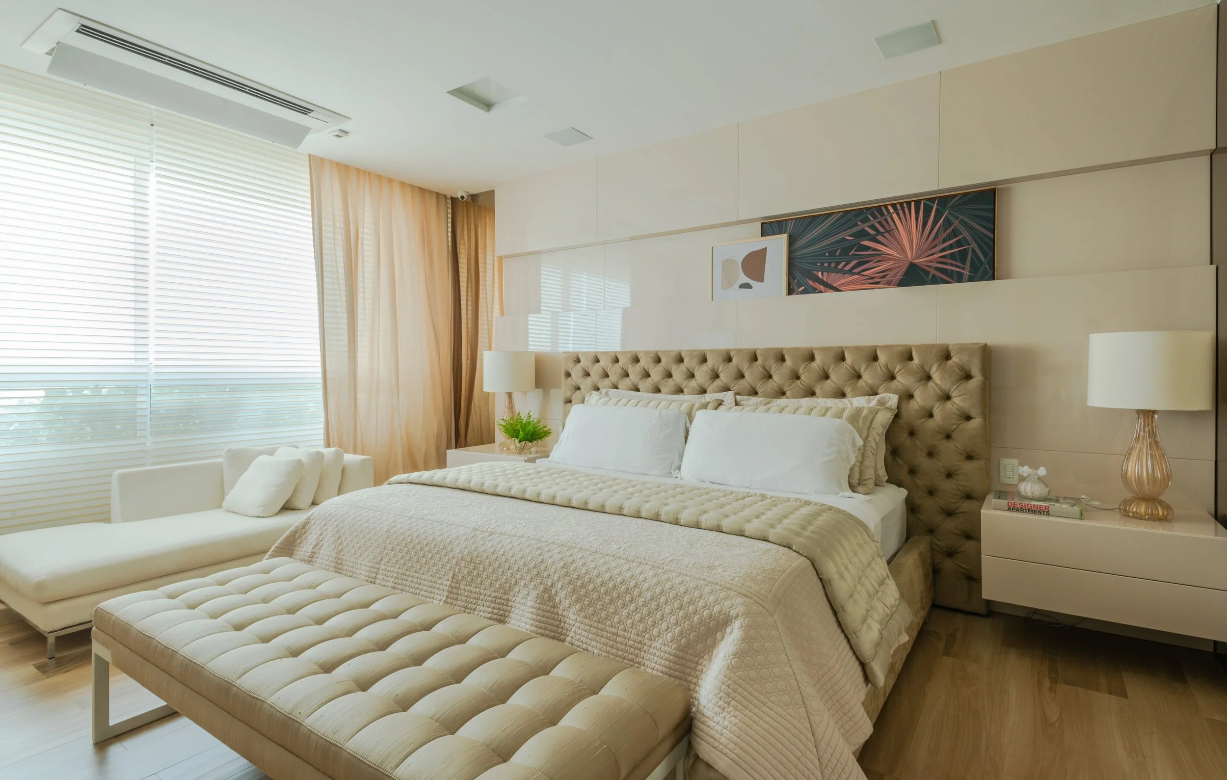 A modern bedroom with a large tufted beige headboard, beige and white bedding, two matching bedside lamps, a cream chaise lounge, and a window with blinds and beige curtains.
