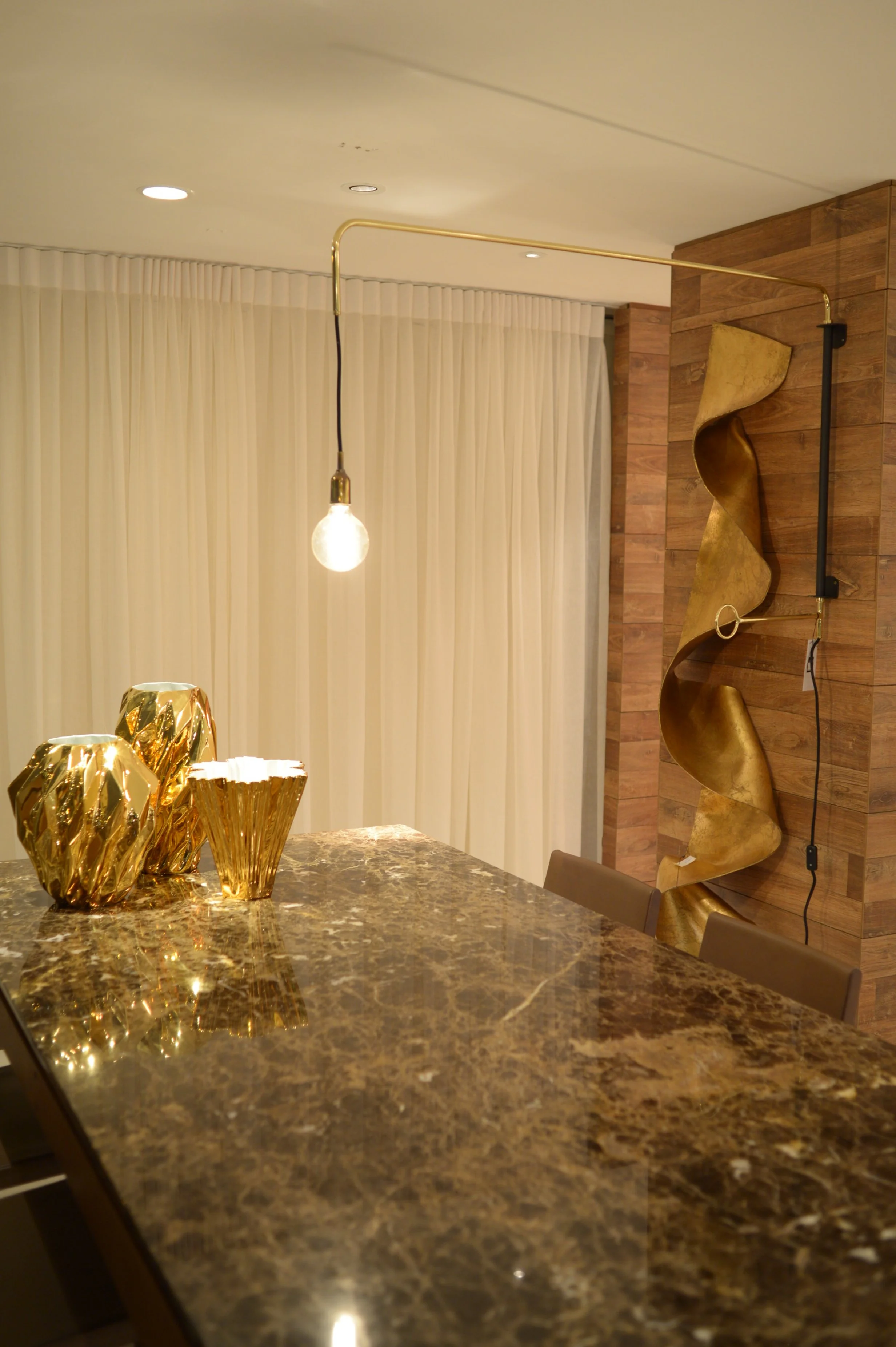Modern dining room with a dark marble table, decorative gold vases, a pendant light, a wooden accent wall with gold sculpture, and cream curtains.